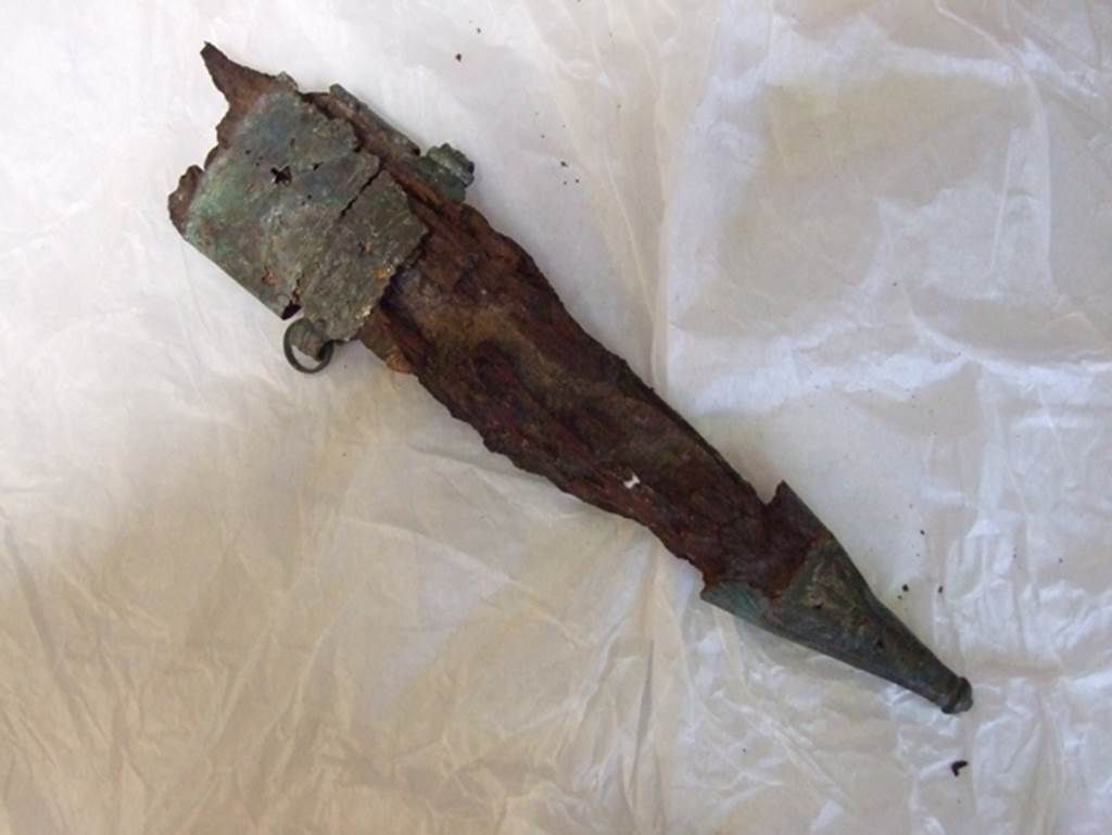 Nola Gate, Pompeii, March 2009. View from above. Large iron knife in a bronze and wooden sheath. 
Found near Porta Nola in 1976. SAP inventory number 17047.
Man no. 5 had on the left a sheathed dagger (fig. 27) and the scabbard (fig. 28) empty of a second smaller blade: at the feet shoes with a nailed sole.
See De Caro S., 1976. Scavi nell’area fuori Porta Nola a Pompei: Cronache Pompeiane V, p. 99, fig. 27.