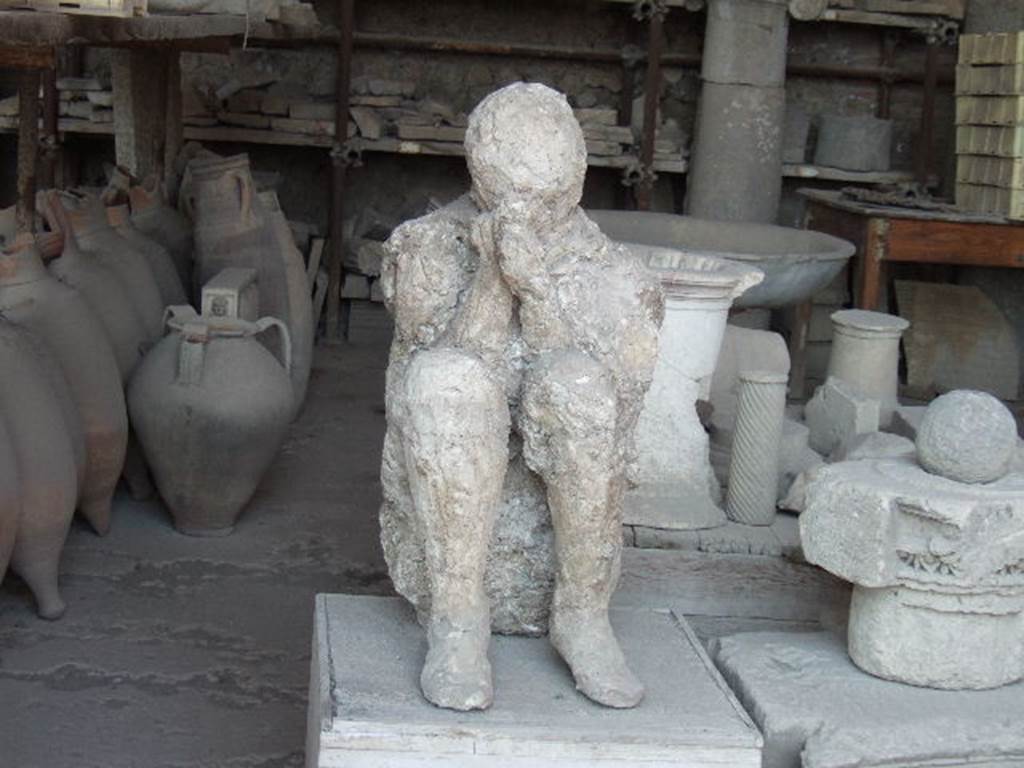 Victim 28.  On display in VII.7.29 Pompeii. May 2006. Plaster cast of body and items in storage.
Two victims were found towards the eastern extremity from the southern portico, and from which plaster-casts could be made because they both lay in the high layer of ash. The one was a beautiful young man's body with agile legs knocked down on the portico floor at the first fall of the ashes.
The other, the most tragically posed, was found leaning against the wall, kneeling, crouching on the ground, in that crouching position that is found in Neolithic tombs, with his head folded forward, bent almost between his knees, for the better protection of himself with his hands and cloak against the inhalation of asphyxiating gases. He had shoes on his feet and wore the typical cloak of the peasant, the “cuculla”, whose flaps he held tight against his mouth.  And in removing the ash under his knees, imprints of overlapping fabrics were noticed, as if the unfortunate man had found comfort in his tragic pose by shielding his knees with the few garments he could arrange. (p.225-6)
For Maiuri’s “Scavo della “Grande Palestra” nel quartiere dell’Anfiteatro, - see Notizie degli Scavi, Anno 1939, Fascicoli 7, 8, 9, on pages 165-238.