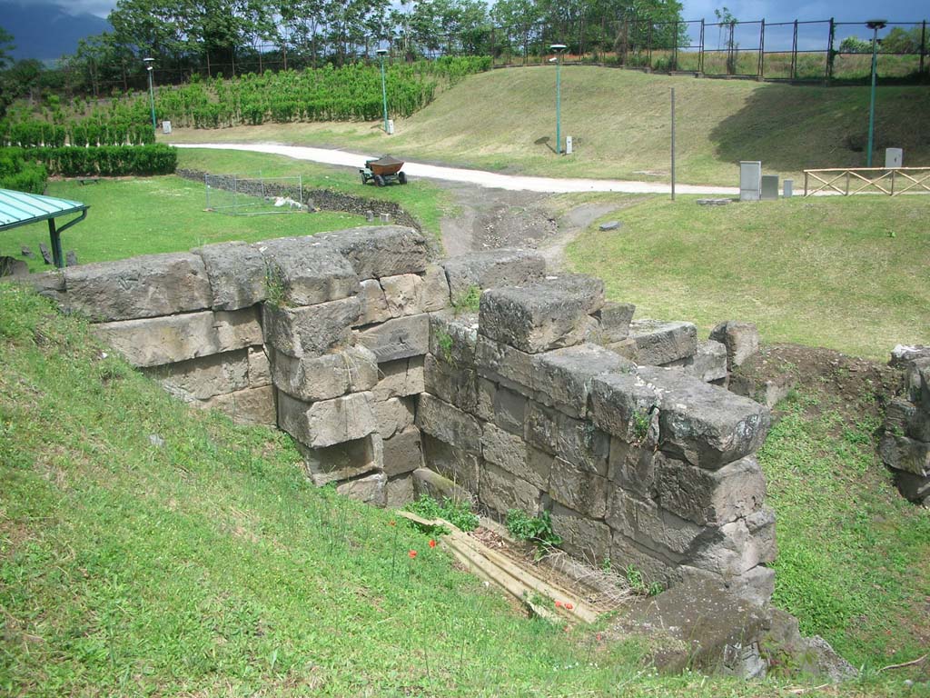 Vesuvian Gate Pompeii. May 2010. Looking north-east from upper area. Photo courtesy of Ivo van der Graaff.
According to Van der Graaff –
“The ornamental emphasis placed on the earliest gates finds a slight variation at the Porta Vesuvio because of its design (see Fig. 3.4).
Its bastions are set slightly aback from the outer curtain. 
This layout created a double bastion on either side of the entrance – a circumstance undoubtedly related to its location in a weak area of the defences. 
The gate was particularly vulnerable to attack because of its position on a natural downward slope in the terrain.
In an effort to strengthen the opening, engineers transformed the northwest tip of the gate into a tower. Today its remains are not immediately evident.
The southern and western facades are buried in the later agger, whereas the northern and eastern flanks are flush with the curtain wall.
Steps recovered in its southern side offered access to the building in its first phase.
The remains represent the only trace of a tower in the first Samnite circuit. Workers probably demolished it after the construction of Tower X slightly further west.”
See Van der Graaff, I. (2018). The Fortifications of Pompeii and Ancient Italy. Routledge, (p.54 and Fig. 3.4).