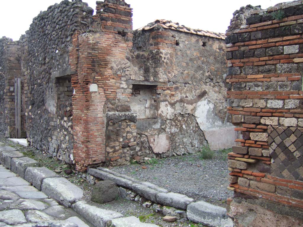 VII.10.2 Pompeii. December 2005. Entrance doorway on Vicolo d’Eumachia, looking north. 
According to Eschebach on the left of the entrance would have been the shop podium or counter, with shelves or steps above it.
See Eschebach, L., 1993. Gebäudeverzeichnis und Stadtplan der antiken Stadt Pompeji. Köln: Böhlau. (p.319)
According to Boyce –
in the north wall is a large rectangular niche (h.0.83, w.0.78, d.0.25, h. above floor 1.0) with a narrow shelf-like projection beside it on the left.
Fiorelli called it “un larario”.
Fiorelli, Descrizione, 273.
See Boyce G. K., 1937. Corpus of the Lararia of Pompeii. Rome: MAAR 14. (p.69, no.306).