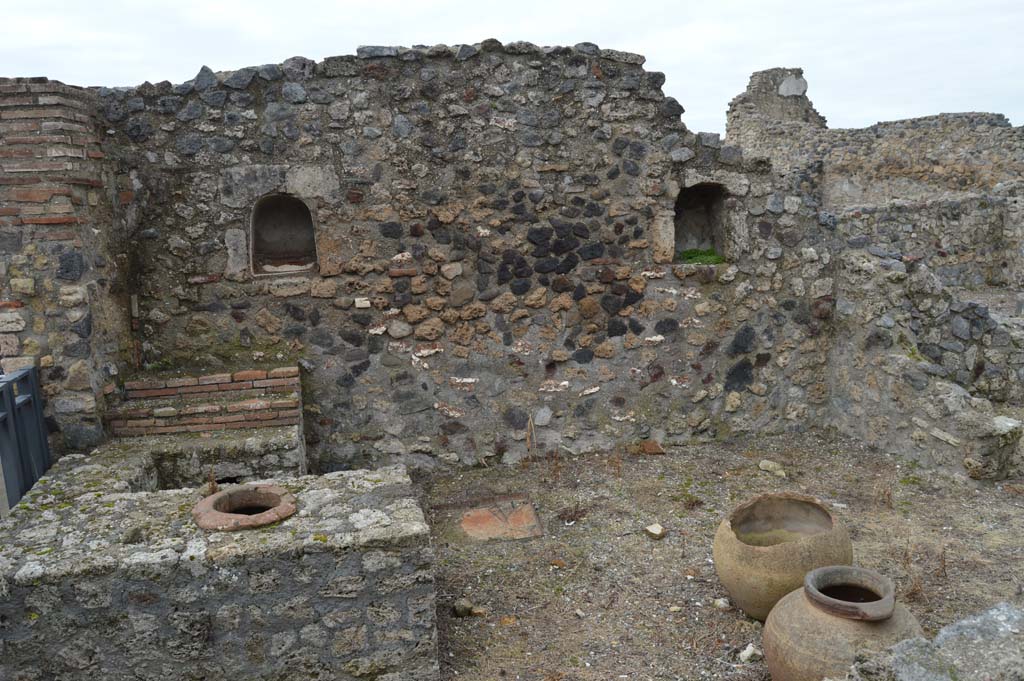 VII.16.7 Pompeii. March 2018.
Looking west towards entrance doorway, on left, counter with display shelves and inset urn/s, and west wall with two niches.
Foto Taylor Lauritsen, ERC Grant 681269 DÉCOR.
According to Boyce –
In the west wall are two arched niches, of practically the same size (h.0.46, w.0.37, d.0.28, h. above floor 1.40, distance apart 2.20), within one of which is a low step – “due nicchia per i Penati”, according to Fiorelli.
See Boyce G. K., 1937. Corpus of the Lararia of Pompeii. Rome: MAAR 14.  (p73, No.337).