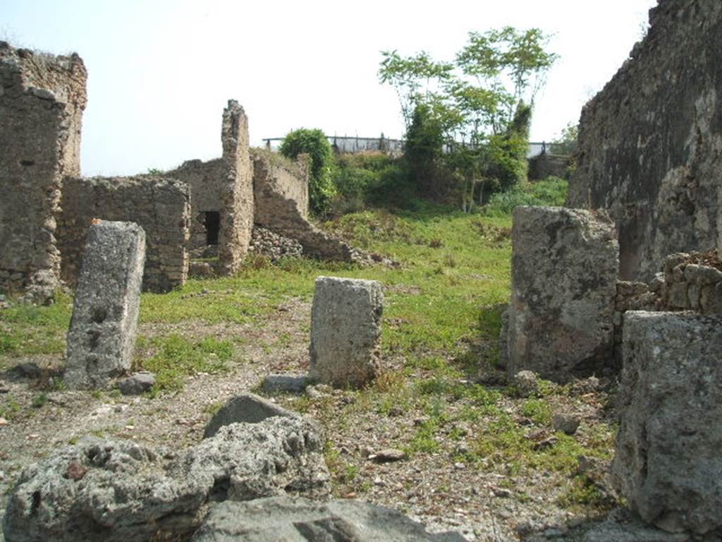 IX.6.4 Pompeii. May 2005. Looking east across site.
The eastern extent of the house can be seen across the centre of the photo, in the middle left.
The pile of stones in the middle centre of the picture, against the north-east wall of the kitchen, would be the site of the hearth, with site of lararium painting above it. 
The wall with a doorway in room “z”, leading to the peristyle of IX.6.5, can be seen in the wall on the left.
According to Boyce – 
In the small open courtyard behind the tablinum in the south wall is an arched niche (h.0.32, w.0.27, d.0.25, h. above floor 2.10).
In its floor was a depression and in the vault of the ceiling a hole; it is called by the Not. Scavi – la piccola nicchia dei Penati.
In the kitchen behind this court, in the north-east corner stands the hearth and on both walls above it is the lararium painting (h.055).
On the north wall is the Genius with cornucopia and patera, pouring a libation upon an altar furnished with offerings, around which  serpent is coiled. On the east wall is a burning altar and on each side of it stands a Lar with rhyton and patera.
See Boyce G. K., 1937. Corpus of the Lararia of Pompeii. Rome: MAAR 14. (p.86, no.429 & 430).
According to Garcia y Garcia, during the night bombing of 16th September 1943, the prothyron, the atrium and four nearby rooms adjoining the south and south-west of this house were hit by a bomb. The pavement and street outside were also damaged. 
Another bomb destroyed a good part of the large room on the east of the house and the perimeter eastern wall of the room on the north-east.
See Garcia y Garcia, L., 2006. Danni di guerra a Pompei. Rome: L’Erma di Bretschneider. (p.153).