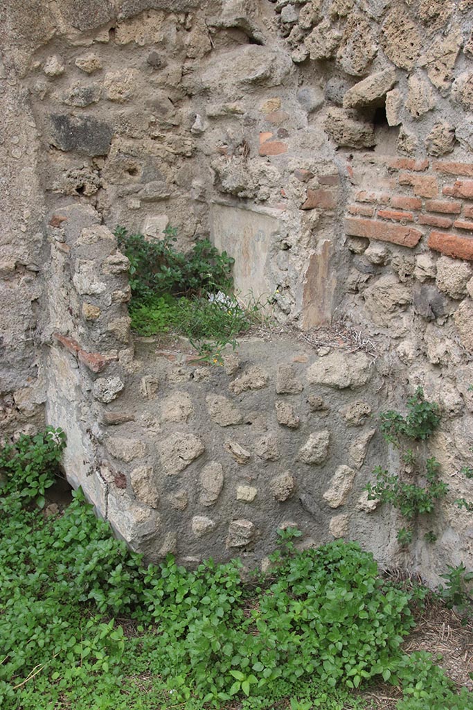 IX.7.12, 14 or 16 (?) Pompeii. October 2024. 
Garden area “e”, lararium in north-west corner. Photo courtesy of Klaus Heese.
According to Boyce –
Against the west wall of the garden stands an aedicula, built upon a square masonry base (0.92 square, h.0.70).
The sides of the base are decorated with green plants painted on the white ground.
Above the base, walls enclose a niche (h.0.42, w.0.52, d.0.62).
In front of the structure stands a small altar (0.24 by 0.23, h.0.80).
The roof (no longer preserved) originally extended out over the altar as well as the niche, and the front of it was adorned with a pediment.
It was supported by two columns, the marks of the bases of which are still to be seen near the corners of the base.
On the walls within the niche is the lararium painting.
On the back wall to the right of an altar, stands the Genius pouring a libation upon it.
To the left of the same altar is a serpent, the head of which was done in stucco relief.
On each of the side walls are painted three figures: from back to front – a plant, a Lar, a man in a white garment holding in his left hand an object resembling a black cup, with a patera in his right hand.
He quotes references – Not. Scavi, 1881, 92-93; Bull. Inst., 1883, 146.
In Not. Scavi, it is described as House, only partly excavated – “and corresponds to, after the sixth doorway, west side, counting from the north-west”.
In Bull. Inst, it is described as “West side, first entrance counting from the south, garden in part excavated.”
See Boyce G. K., 1937. Corpus of the Lararia of Pompeii. Rome: MAAR 14. (p.89, no.445).