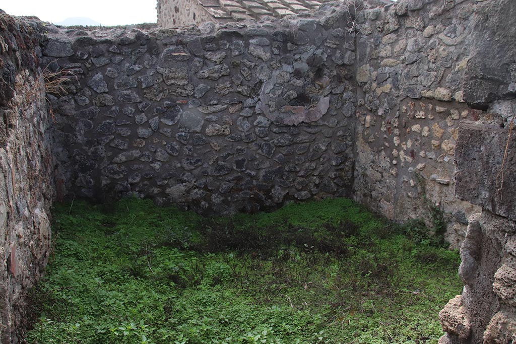 IX.7.19 Pompeii. October 2024. Room k, looking south through doorway in corridor f. Photo courtesy of Klaus Heese.
The east wall and south-east corner are on the left, presumably the site of the lararium painting, but nothing now remains.
According to Boyce, a small room, room k, located between kitchen and garden on the south side of the house, was originally part of the garden.
This small room had a doorway into the kitchen. 
Then the room was separated from the garden and the doorway to the kitchen was bricked-up.
On the east wall of this new room was a lararium, with a Genius standing to the right of a tripod.
On the other side of the tripod stood the tibicen. Below and to the right, ran a camillus. 
Below this were two serpents, gliding, one from each side, amongst plants towards an altar.
Between this painting and the south-east corner of the room were painted, two pots, a large bottle, sausages, a calf’s head, ribs of pork on a spit, and a phallus.
He references – Not. Scavi, 1880, 491; Bull. Inst., 1883, 82.
See Boyce G. K., 1937. Corpus of the Lararia of Pompeii. Rome: MAAR 14. (p.88, no.438) 
According to NdS, the were also two Lares:
To right in the atrium there is a room communicating with the back room of the following taberna, and the access to the kitchen with the latrine, and to a cell, where the lararium painting is divided into two strips; in the lower part there are the two serpents, which rush towards the altar with the offerings, and in the upper part the sacrificing Genius Familiaris, with tibicen and a camillus between the two Lares.
See Notizie degli Scavi di Antichità, 1880, p. 491.
Fröhlich notes: Boyce does not mention Lares.
Boyce 438 (IX 7, 19) describes the Genius with Camillus and Tibicen grouped around the altar in the usual way, but without the Lares. 
See Fröhlich, T., 1991. Lararien und Fassadenbilder in den Vesuvstädten. Mainz: von Zabern. (p.112).
See Giacobello, F., 2008. Larari Pompeiani: Iconografia e culto dei Lari in ambito domestico. Milano: LED Edizioni, (p.209, no.106)