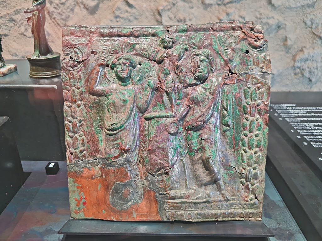 Civita Giuliana, Imperial Villa, March 2024. Silver and bronze panel from the Isiac lararium. Photo courtesy of Giuseppe Ciaramella.
Silver plated bronze panel, with embossed Isiac triad (1st century AD). (Laminetta con triade isiaca a rilievo, bronzo argentato, I sec. d.C)
On display in exhibition in Palaestra entitled – “L’altra Pompei, vite comuni all’ombra del Vesuvio”.