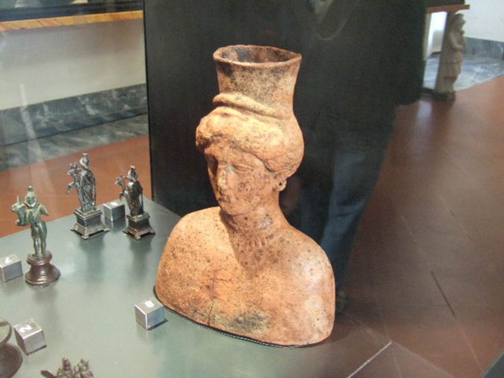 I.2.20 Pompeii. Bust of Isis found in Caupona at I.2.20. Now in Naples Archaeological Museum. According to Boyce, a terracotta bust of a woman with a modius upon her head and adorned with a necklace was found in the niche.
Also found in the niche, were: A bronze equestrian statuette of a helmeted warrior, holding a spear in his right hand. A bronze statuette of Diana, holding a bow in her left hand, she was drawing an arrow from the quiver on her shoulder with the right hand. A terracotta statuette of a woman, fully clothed, seated and holding a child in swaddling clothes.
A terracotta object in the form of a kind of cradle, within which lies the half-bust of a child with a bulla about its neck. A green-glazed clay head representing Medusa. A marble Bacchic head from a herm.
See Boyce G. K., 1937. Corpus of the Lararia of Pompeii. Rome: MAAR 14. (p.23).