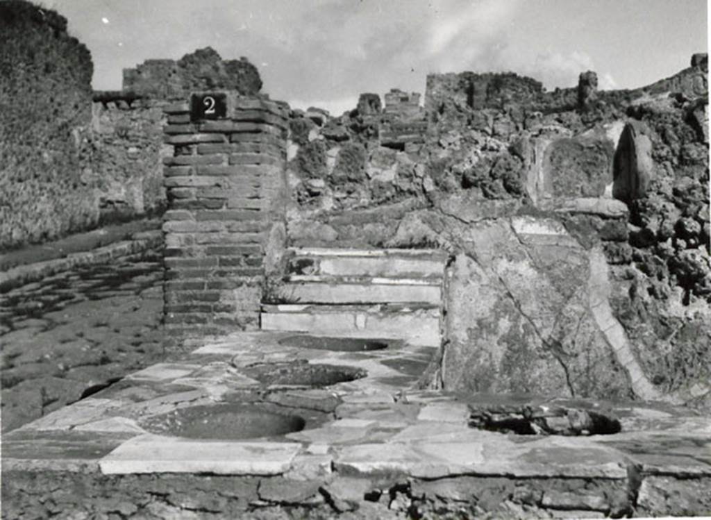 I.3.2 Pompeii. 1935 photograph by Tatiana Warscher. Looking north along west side of the counter showing four embedded dolia, with Via Stabiana on the left. The niche, as mentioned by Boyce above, can be seen in the north wall. According to Warscher, quoting Fiorelli – “I.3.2,  Bottega con podio, in cui stanno infisse quattro grandi urne di terracotta, avendo a destra una gradinata. Nel muro nord si trovano i resti della nicchia dei penati”.
See Warscher, T, 1935: Codex Topographicus Pompejanus, Regio I, 3: (no. 3), Rome, DAIR, whose copyright it remains.