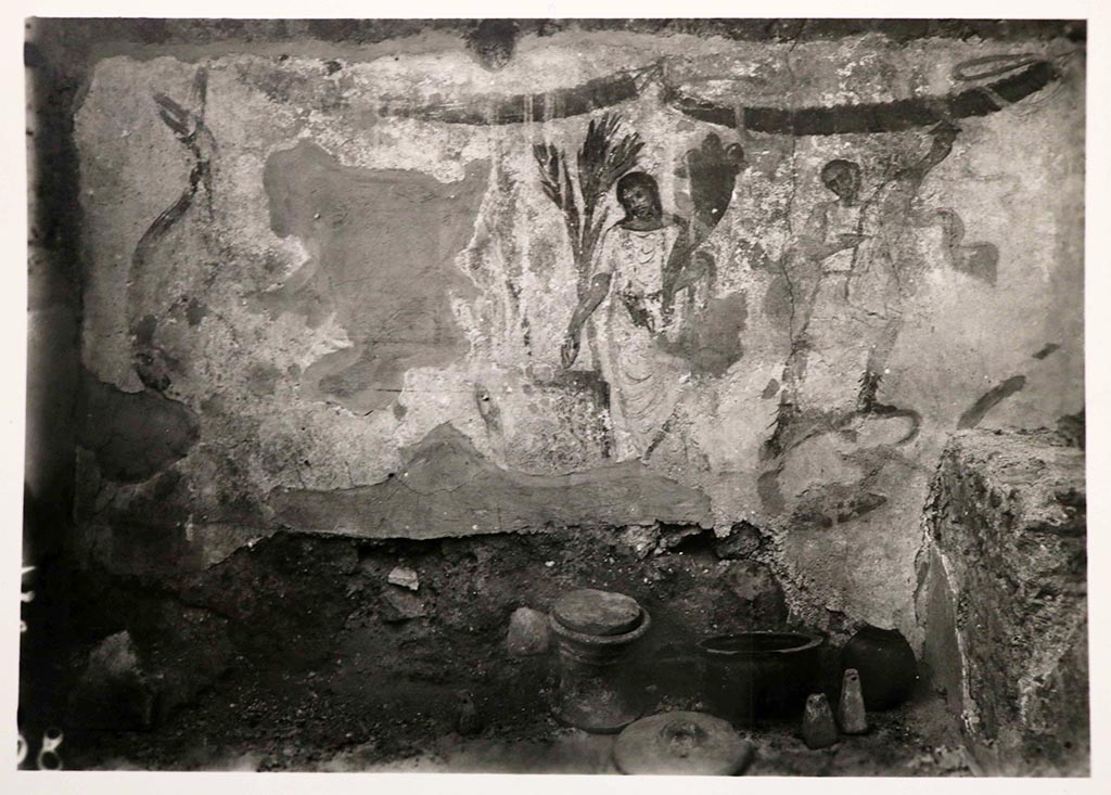 I.7.18 Pompeii. Lararium painting, north and west wall in kitchen, found above the hearth, on which were also found furnishings, ceramics and weights.  
See Giacobello, F., 2008. Larari Pompeiani: Iconografia e culto dei Lari in ambito domestico. Milano: LED Edizioni, (p.138, no.11)
According to Fröhlich this was painted on a white-ground wall surface. The picture on the north wall shows the sacrificing genius flanked by two lars at the round altar, behind which stand two palm bushes. Garlands hang from the upper edge of the picture.
To the right of the head and arm of the right-hand Laren, the corresponding body parts of an older Laren show through under the current white plaster. Remnants of brown paint on the left of this scene are probably from a coiled snake stretching its head up towards the corner of the room. The faint traces of paint on the plaster stucco on the adjoining west wall suggest that an analogous snake was depicted here. If this is the case, the two snakes may have flanked an altar painted in the corner of the room, cf. L66 (VI 9,6/7).
See Fröhlich, T., 1991. Lararien und Fassadenbilder in den Vesuvstädten. Mainz: von Zabern, (p. 252, L7, taf. 25.1).