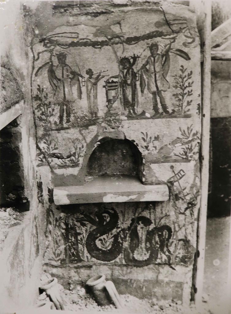 I.8.14 Pompeii. 1938. 
Lararium painting and niche in north-east corner of courtyard 7, near to the kitchen (10), no longer conserved.
According to Giacobello, this is found in the north-east corner of viridarium 7 of I.8.14. Fröhlich had located this in insula I.8 but could not assign it to a specific house.
Above the niche is the painting showing two large Lares, a smaller togated Genius with a cornucopia and sacrificing on a round altar while on the other side of the altar a tibicen is playing the double flutes.
At the sides of the niche are two birds in shrubs.
Below the niche is a serpent approaching an altar.
See Giacobello, F., 2008. Larari Pompeiani: Iconografia e culto dei Lari in ambito domestico. Milano: LED Edizioni. (p.141, no.13).
Fröhlich describes a wall surface painted on a white background with an arched niche. In the uppermost zone, the sacrificing Genius stands on the right and Tibicen on the left at the round altar. The scene is flanked by two Lares, next to which a laurel bush stands on each edge of the picture. A garland is hung on the upper edge. The arched niche is recessed into the narrow, middle zone. On both sides of the niche, a peacock appears between small plants. In the lowest zone, a snake moves between marsh plants to the left towards a round altar. At the top right, a skewer with three pieces of meat is painted. In the right half, the remains of an older snake painting can be seen under the plaster, which may also include the adjacent zone with small plants, set off in color and by a plaster edge, to which a dark base is attached below. On the wall to the left, a pig's head is painted below and a ham above it. It is not known which phase these eating still lifes belong to. In any case, they suggest that the painting was located in a kitchen. 
See Fröhlich, T., 1991. Lararien und Fassadenbilder in den Vesuvstädten. Mainz: von Zabern, See Fröhlich, T., 1991. Lararien und Fassadenbilder in den Vesuvstädten. Mainz: von Zabern, p. 254, L11, Taf. 26,2.