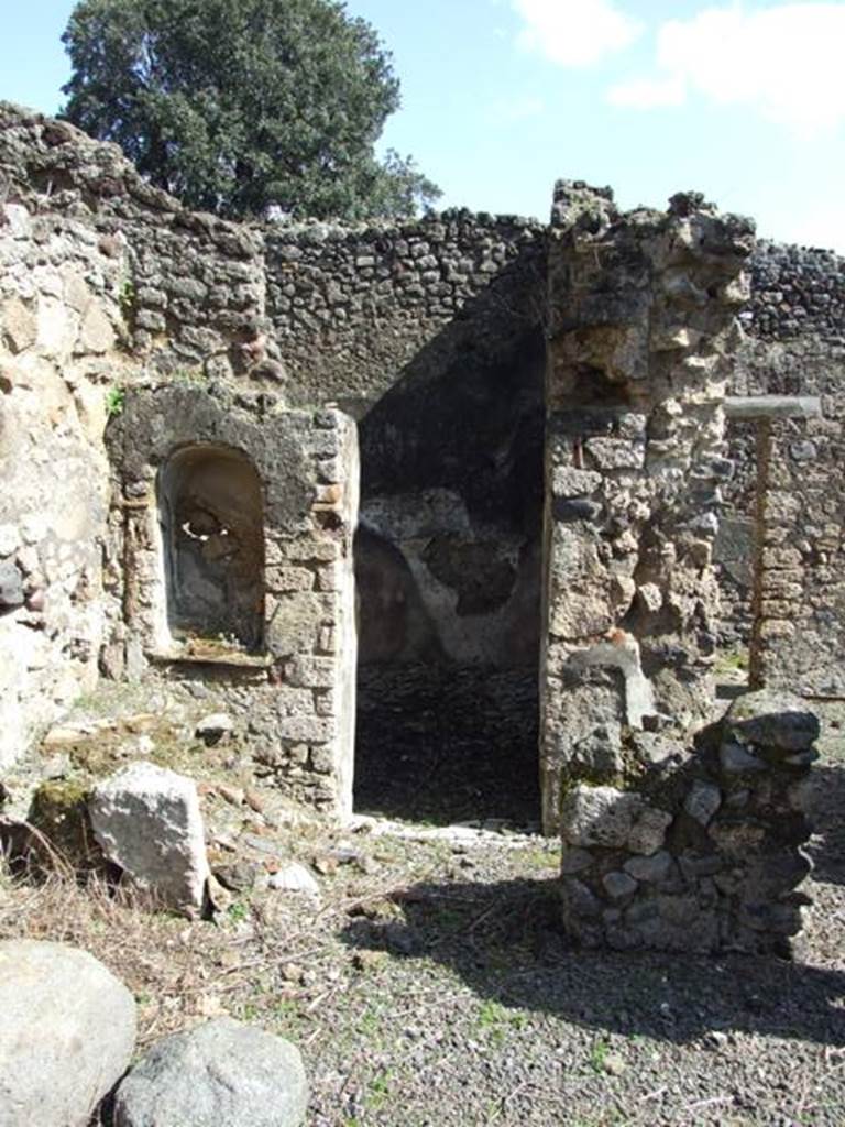 V.4.9 Pompeii. March 2009. Kitchen on east side of atrium. Looking east. According to Boyce, on the south-east corner of the impluvium was a square column (see remains on right of photo). This column closed off part of the east side of the impluvium to provide a small area as a kitchen. In the west side of this column, facing the impluvium, was a niche with slightly arched ceiling and projecting floor. Above and to the right of the niche was a yellow laurel wreath with pendent taeniae hung from a nail – all in stucco relief (see photo below). Within the niche were found – an iron knife with bronze handle, an iron hammer, and a bronze rosette. On the same side of the column, but higher, was another larger rectangular niche, probably for storage purposes.
See Boyce G. K., 1937. Corpus of the Lararia of Pompeii. Rome: MAAR 14. (p.41, no.124, Pl.4,5) 
Hardly anything remains of the column today, and neither niche.
In the north-east corner of the kitchen stood a small hearth and above it in the east wall was a high arched niche (see above photo, on the left). Its walls were coated with white stucco. On its floor stood a masonry block, apparently for use as an altar, behind it a step was built against the rear wall. In the centre of the step was a mark left by an oval object which had stood there, probably a statuette. Another altar, terracotta and cylindrical, was found within the niche.
See Boyce G. K., 1937. Corpus of the Lararia of Pompeii. Rome: MAAR 14. (p.41, no.125, Pl.4,5)