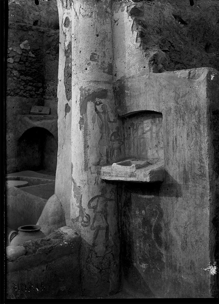 V.4.b Pompeii. 1931. Lararium found on the east wall of the atrium, near the kitchen door.
DAIR 31.2876. Photo © Deutsches Archäologisches Institut, Abteilung Rom, Arkiv. 
A heavy shelf with a low raised area for a built-in altar projects from a niche with a slightly arched ceiling.
Within the niche was a crudely done lararium painting on a yellow background.
Above and to the sides hung garlands.
On the back wall of the niche the Genius pours a libation upon a cylindrical altar, on the opposite side of which stands the tibicen. 
On the wall outside the niche is a Lar on each side, the one on the right holding rhyton and situla, that on the left rhyton and a cup like a skyphos. 
On the left side wall of the niche, between the Lar on that side and the back wall of the niche, is the popa leading a hog. 
Below the level of the niche on the left side, is Mercury with petasos, caduceus and purse; behind him, suspended, two crescent-shaped objects. 
These three figures on the left side are painted on the column of the peristyle in the south-east corner of the garden. 
Immediately below the niche a crested serpent (two serpents according to Sogliano in the Not. Scavi, one according to Mau in the Rom. Mitt.) glides to the left among plants.
See Boyce G. K., 1937. Corpus of the Lararia of Pompeii. Rome: MAAR 14. (p.42, no.129, Pl 13,1)
See Jashemski, W. F., 1993. The Gardens of Pompeii, Volume II: Appendices. New York: Caratzas, p. 117
See Giacobello, F., 2008. Larari Pompeiani: Iconografia e culto dei Lari in ambito domestico. Milano: LED Edizioni, p.168.