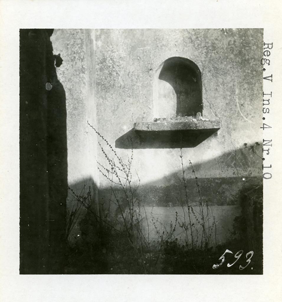 V.4.c Pompeii but shown as V.4.10 on photo, 1937 or earlier, by Tatiana Warscher. Niche lararium (e) in east wall of garden K.
Photo courtesy of American Academy in Rome, Photographic Archive. Warsher collection no. 593.
According to Boyce, this niche was placed at the east end of the corridor leading from the atrium.
The arched niche had a heavy ledge projecting from the wall. 
Upon this ledge was placed an ordinary roof tile, the raised edges of which, when covered with stucco would look like an altar.
The rear wall was painted blue or green, and painted with plants.
Boyce was not sure if this was a Lararium, but he thought as there was no other Lararium in the house, it may possibly have been one.
He said that Sogliano attached religious importance to a painting of Mercury found on the east wall of the atrium. 
See Notizie degli Scavi di Antichità, 1905, p. 131.
See Boyce G. K., 1937. Corpus of the Lararia of Pompeii. Rome: MAAR 14. (p.42, no.130, Pl. 3,1 - which is this photo by Warscher)
In a note on page 42, Boyce added that a collection of items were found, perhaps, gathered together for flight.
Included were a glazed terracotta statuette of Harpocrates and a small terracotta altar.
See Bullettino dell’Instituto di Corrispondenza Archeologica (DAIR), xvi, 1901, 364.