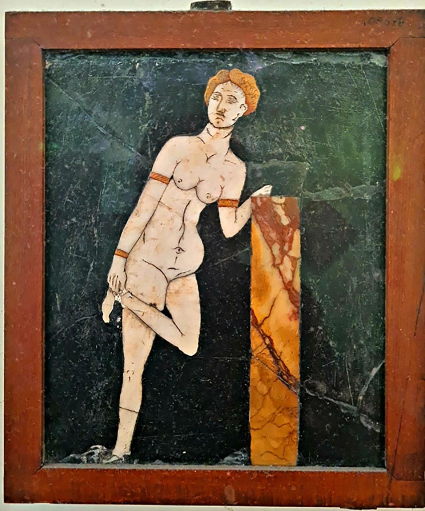 .2.10 Pompeii. Found in triclinium. Photo courtesy of Giuseppe Ciaramella. September 2024.
Now in Naples Archaeological Museum.  Inventory number: 109678.
According to the information card –
Venus. Slate wall panel with polychrome marble inlay work.
The theme shows Venus with her hair tied back, with bracelets on her wrists and arms. 
She leans with her right elbow on a small column as she unties a sandal. (Third quarter of the Ist century AD.)
(Venere. Panello parietale di ardesia a intarsi marmorei policromi.
Il soggetto rappresenta une Venere dai capelli legati, con bracciali ai poisi e alle braccia, la quale, poggiandosi con il gomito destro a un pilastrino, si slaccia un sandalo. Terzo quarto del I secolo d.C.)