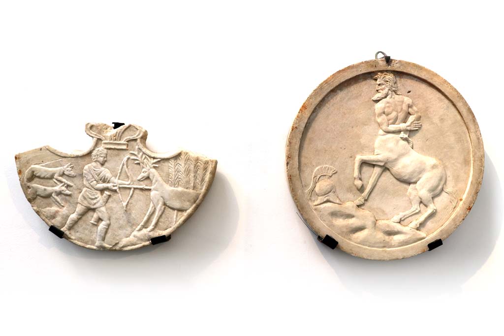 I.4.25 Pompeii. February 2021. On left, pelta hung between columns in the portico, showing scenes of Hercules’ life.
On right, Oscilla with centaur from VI.16.7. On display and photographed in Antiquarium.
Photo courtesy of Fabien Bièvre-Perrin (CC BY-NC-SA).