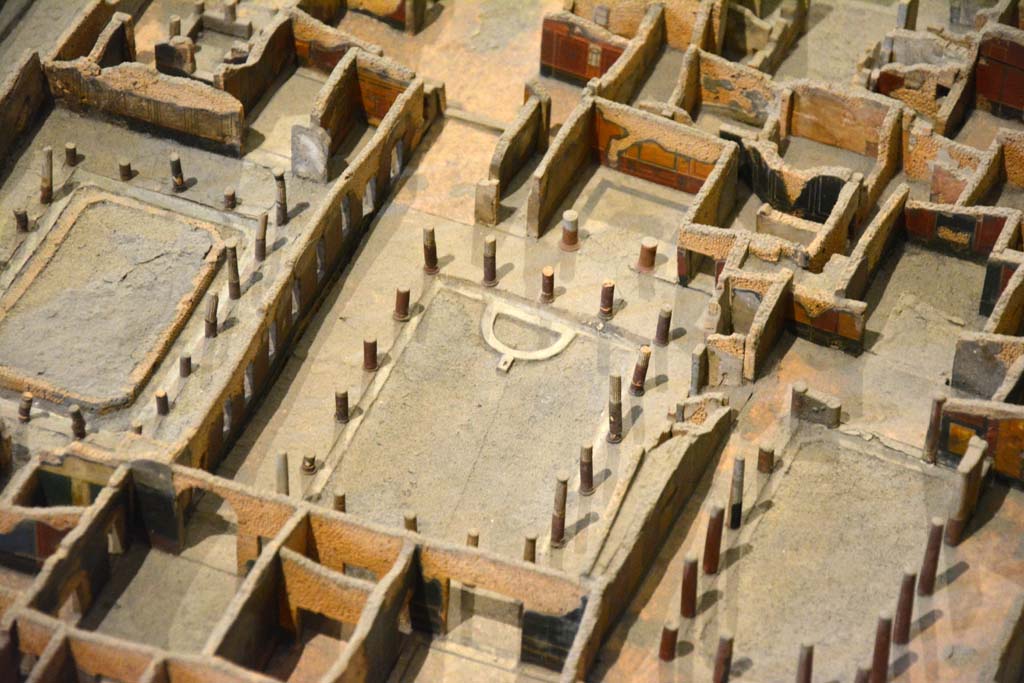 I.4.25 Pompeii. May 2019. Detail from model in Naples Archaeological Museum.
Looking south-west across Middle Peristyle 17, in centre. 
Foto Tobias Busen, ERC Grant 681269 DÉCOR
According to Jashemski –
“The centre garden, (the Middle Peristyle), was surrounded by a portico supported by seventeen columns made of brick and Sarno limestone and faced with stucco. The upper parts were fluted, the bottoms left plain and painted red. There was a gutter around the edge of the garden. 
At the west end of the garden was a semi-circular basin around which were arranged six spectacular bronze animals found in 1861.  …………………………
In February 1926, when restoring the garden in this peristyle, Maiuri discovered a large rectangular pool (3.50m wide x 10.80m long x 1.40m deep). On the west side two high steps (0.45m and 0.40m) led down to the pool. The rough foundations of the much smaller semi-circular pool had been built on these steps and on the bottom of the large pool. The large pool had then been filled with old building material and soil. This was probably at the time that the house with the entrance on the Via dell’Abbondanza was joined to this house, probably after the earthquake. The cistern opening in the south-west corner of the garden had a lava puteal; the one in the east portico, no puteal.”
See Jashemski, W. F., 1993. The Gardens of Pompeii, Volume II: Appendices. New York: Caratzas. (p.29-30).