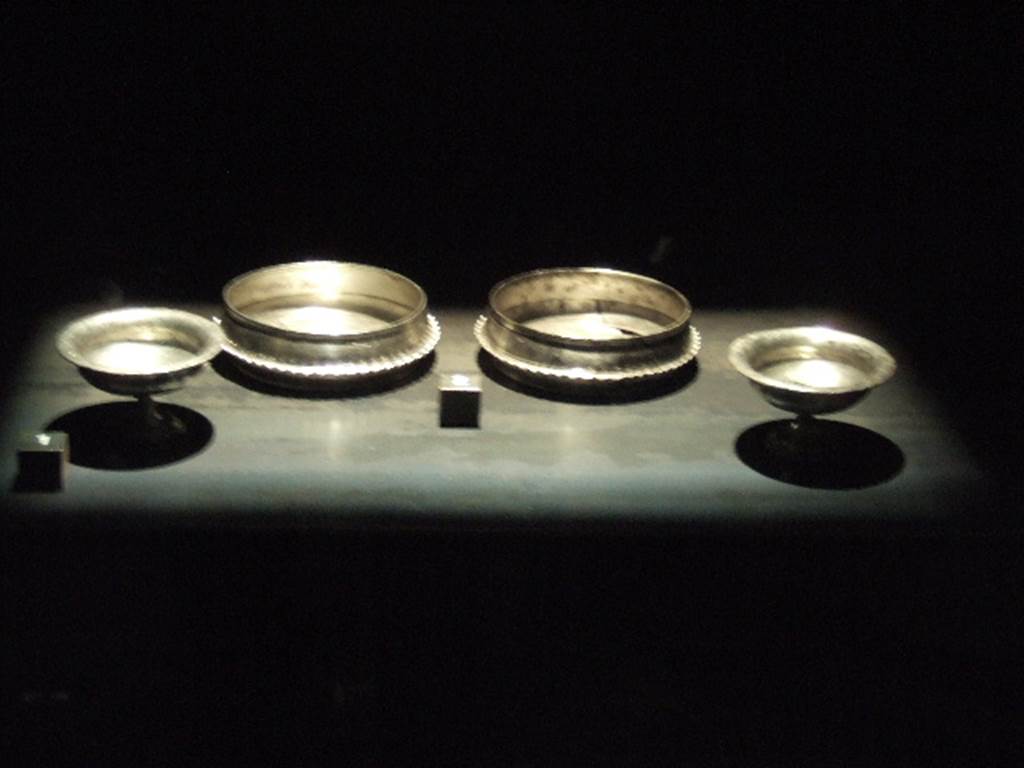 Silver found against west wall of atrium of I.6.11. 
At the edges are two goblets each in the form of a chalice.
At the centre are two bowls (ciotole) with the upper part with high edges decorated with concentric circles parallel to the rim.
Below this is a lip with an undulating ribbon pattern which separates the upper and lower parts . 
Now in Naples Archaeological Museum. 
Bowls:    inventory numbers 144804 and 144805.
Goblets: inventory numbers 144806 and 144807. 
See Guzzo, P. (A cura di), 2006. Argenti a Pompei. Milano, Electa. p.141.