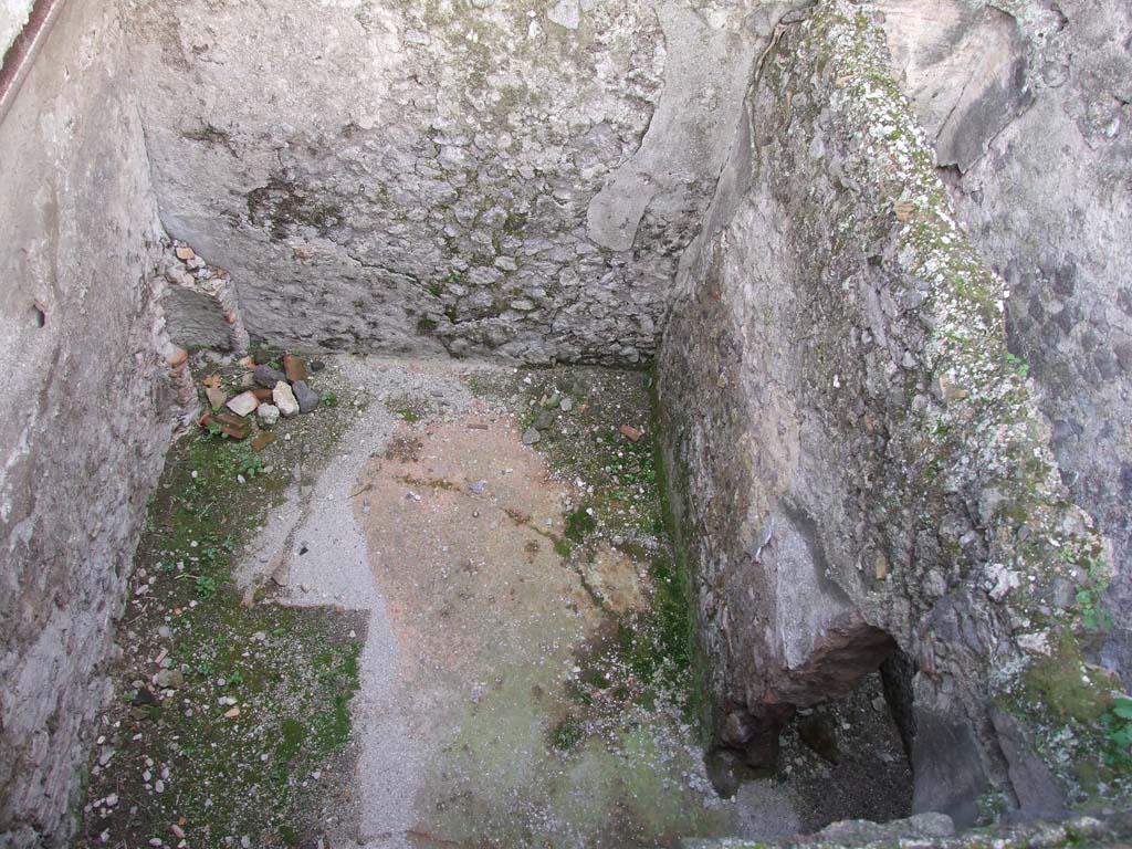 I.7.1 Pompeii. December 2006. 
Lower level room, with corridor, on right, and remains of masonry stove, on left.
According to NdS –
“The corridor and the room must have belonged to a bath in a more ancient period: the corridor still retains its decoration with a beautiful red background, with a small doorway with a round arch and a circular opening walled-up in the rear renovations, the room has clear traces of the rib of a stuccoed vault, elements, one or the other, completely foreign to the rustic character of all the other basement areas of this house; in the corner of the room you can see the remains of a brick stove that could also be used to heat the room and a hole on the floor for water to escape.
Once the bath room was abolished, the simple wooden ceiling was left on the room, and to ventilate and illuminate it, the ingenious expedient of leaving the spaces empty between beams, on the top of the wall, was resorted to. This was in order to obtain as many glimmers of light and air from the void between two beams”.
(Il corridoio e la stanza debbono avere appartenuto in un periodo piu antico ad un bagno: il corridoio conserva infatti ancora la sua decorazione a bel fondo rosso, con una porticina ad archetto tondo ed uno spiraglio circolare murati nei rifacimenti posteriori, la stanza ha chiare tracce della centina di una volta stuccata, elementi l’uno o l’altro del tutto estranei al carattere rustico di tutti gli altri sotterranei di questa abitazione; nell’angolo della stanza si osservano i resti di un fornello in mattone che poteva anche servire al riscaldamento dell’ambiente ed un foro al piano del pavimento per la fuoruscita dell’acqua.
Abolito il bagno si lascio il semplice solaio in legno sulla stanza, e per arieggiarla ed illuminarla si ricorse all’ingegnoso espediente di lasciare sul collo del muro gli spazi vuoti fra trave e trave in modo da ricavare dal vacuo tra due travi altrettanti spiragli di luce e di aria.)
See Notizie degli Scavi, 1929, (p.390-391).