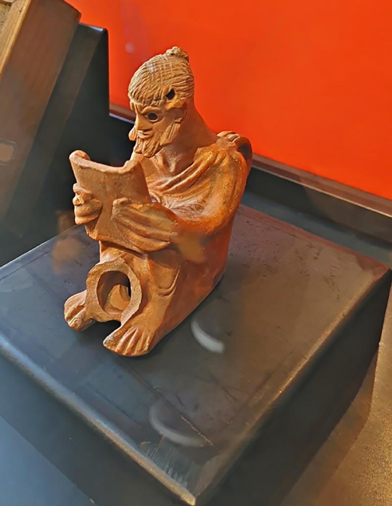 I.8.14 Pompeii. March 2024. 
No.2 on information card – Oil lamp with an intellectual thinker (Ist century AD). (Lucerna plastica con intellettuale (I sec. d.C).
Photo courtesy of Giuseppe Ciaramella.
On display in exhibition in Palaestra entitled – “L’altra Pompei, vite comuni all’ombra del Vesuvio”.