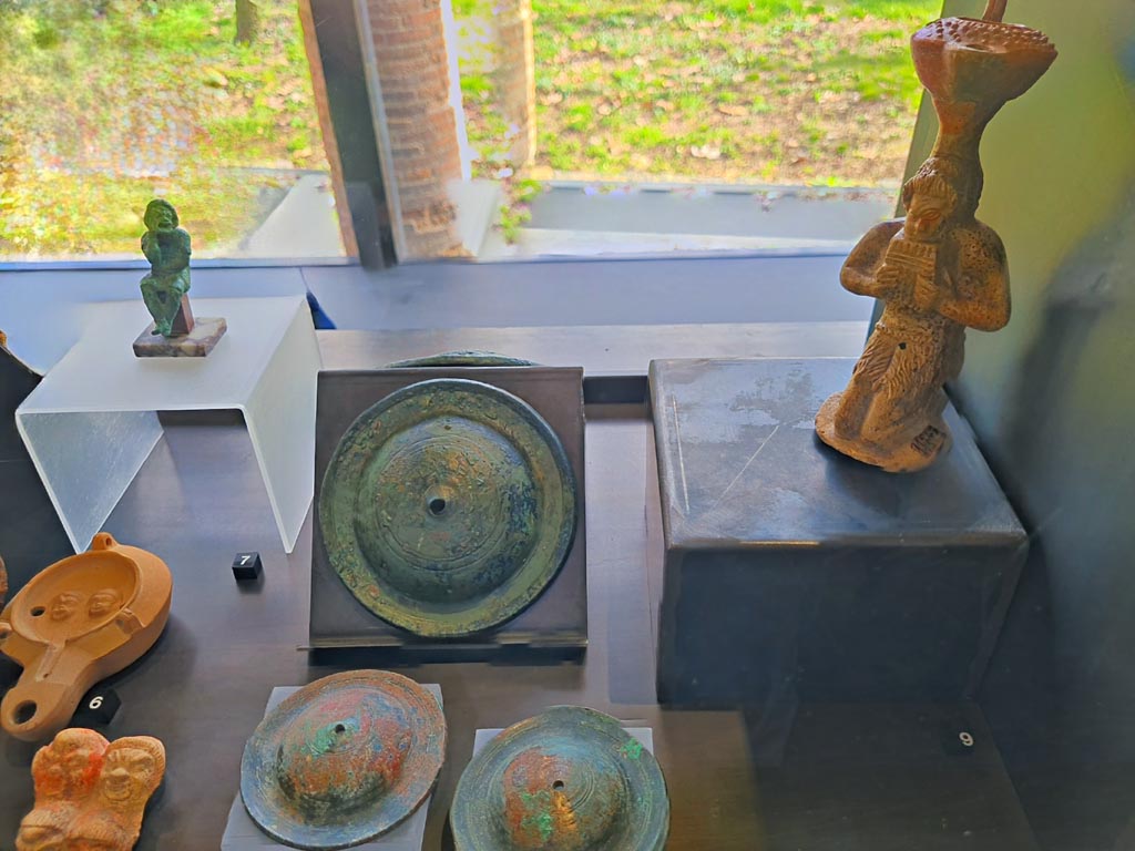 I.8.17 Pompeii. March 2024. No.7 – Bronze statuette of an actor (top left) (1st century AD). Photo courtesy of Giuseppe Ciaramella.
On display in cabinet in exhibition in Palaestra entitled – “L’altra Pompei, vite comuni all’ombra del Vesuvio”.
No.6 – Oil lamp with theatre masks (1st century AD), from I.9.9.
No.7 – Bronze statuette of an actor (1st century AD), from I.8.17.
No.8 – Bronze cymbals (1st century AD), provenance not known but from Pompeii. 
No.9 - Oil lamp with Pan (1st century AD), from I.13.2.