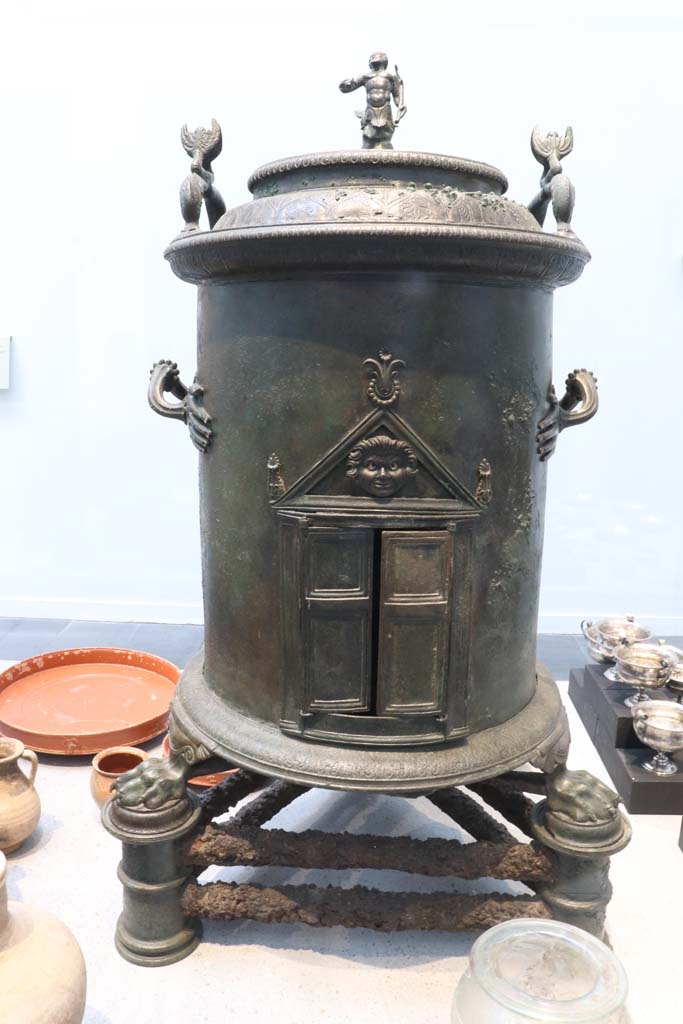 VIII.1.4 Pompeii. February 2021.
Bronze cylindrical food warmer found under the stairs in the servant’s quarters of I.8.17.
On display in Antiquarium. Photo courtesy of Fabien Bièvre-Perrin (CC BY-NC-SA).
According to the Musée Maillol, the Romans created a range of richly decorated devices, usually in bronze, for storing hot water during triclinium banquets. 
The hollow cylinder of the latter rests on an iron tripod. 
The feet are in the form of lion's paws, the handles end in small hands. 
A small temple with a Gorgon in the tympanum can be seen in the opening.
Two dolphins and a Triton decorate the lid.
See Le Musée Maillol : Pompéi - Un art de vivre, l’exposition du 21 septembre 2011 au 12 février 2012. Communiqué de presse.
Parco Archeologico Pompei, inventory number 6798.