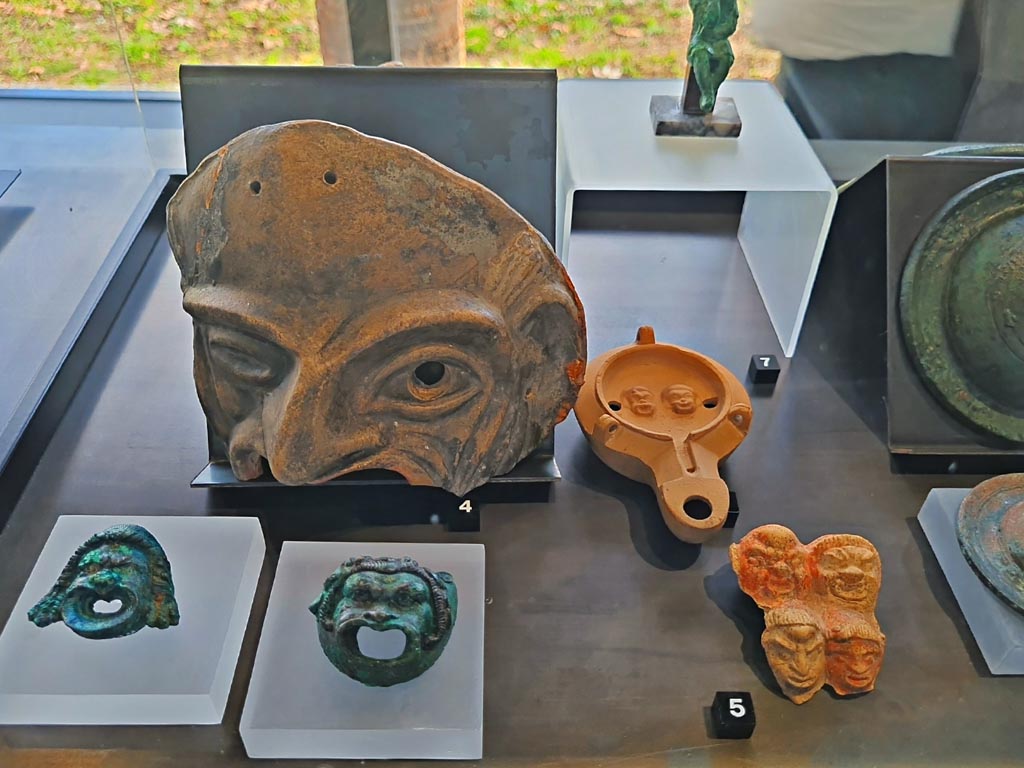 I.9.13/14 Pompeii. March 2024. No.5 – Plaque with theatre masks, (Ist century BC – Ist century AD). Photo courtesy of Giuseppe Ciaramella.
On display in exhibition in Palaestra entitled – “L’altra Pompei, vite comuni all’ombra del Vesuvio”.
No.3, lower left – Bronze wall sconces with theatre masks, provenance not known, but from Pompeii. (Ist century AD).
No.4 – Terracotta theatre masks Pompeii – provenance not known, but from Pompeii. (Ist century AD). 
No.6 (number hidden, but beneath No.7) – Oil lamp with theatre masks, from I.9.9.