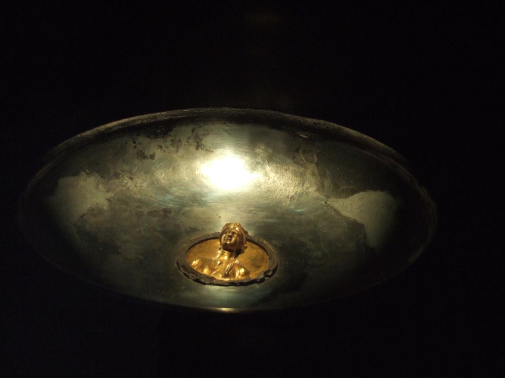 I.10.4 Pompeii. Silver phiale or flat drinking vessel with gold emblem in centre. 
The cup appears to have been used for display and a stand for this was found also.
Part of the 115 pieces of Silver found in a chest in the underground storerooms. 
Now in Naples Archaeological Museum. Inventory number 145544.
See Guzzo, P. (A cura di), 2006. Argenti a Pompei. Milano, Electa. p.220-1.