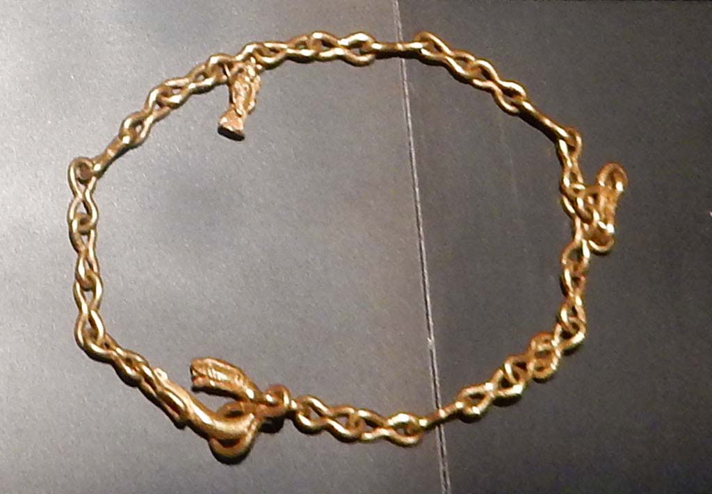1.10.7 Pompeii, June 2019. 
Heavy gold chain with figure-of-eight links, snake head fastening and pendant featuring a statuette of Isis Fortuna, holding a rudder and cornucopia.
This was one of the jewels found in the atrium and in the tablinum.
On display in Palestra exhibition of jewellery entitled “Vanity, storie di gioielli dalle Cicladi a Pompei”, from 10th May 2019 to 5th August 2019.
SAP inventory number 5413. Photo courtesy of Buzz Ferebee.
See Guzzo, P.G. ed. (2003): Tales from an eruption – (Civale, A: The House of the Craftsman). Milan, Electa; (p.140-42)