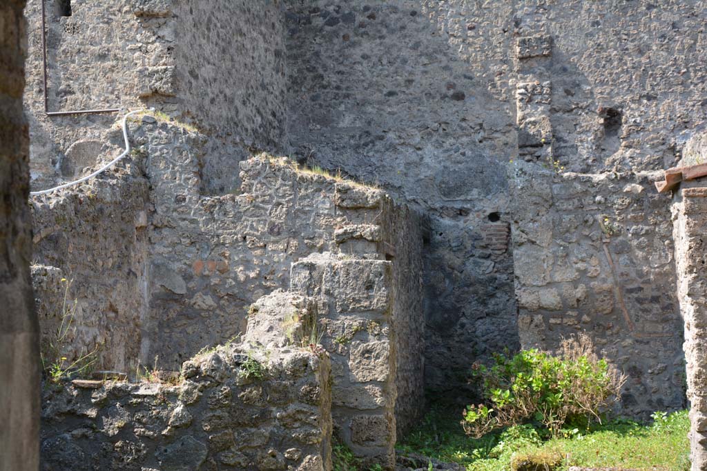 I.10.18 Pompeii. April 2017. Looking south-west from entrance fauces. Photo courtesy Adrian Hielscher.
“Along the south side (of the atrium 1) which was the wall next to the rustic quarters of the Casa del Menandro, a block of limestone was found, which would have been used as reinforcement for the wooden stairs that led up to the upper floor, of which one could see the outline in the plaster.
The “understairs”, closed towards the atrium by a wooden partition, had the two brick walls covered in simple white plaster.
On the west side of the atrium opened, respectively to the right and to the left, 
- a large doorway opening into a room all open to the west (no.6) and defended only by a low wall, covered by canopy/roof,
- and with another room (no.7) of elongated trapezoidal shape, whose south wall, slanting, marks a tilt of the perimeter wall.
In the room to the right, (no.6) there is no trace of the original flat roof cover other than the two large holes of the lintel, there is, on the left side, a recess in the wall, to which a pile of lime had been attached and on the right side were the two limestone blocks, supporting an attached ladder. The flooring was of a simple floor of beaten material; the walls were covered with rustic white plaster, which did not have any decoration.
From here we pass by means of a door rather wide, but occupied in part by the circular mouth of the cistern, into a narrow and long area (no.8), uncovered, with a floor of cocciopesto, onto which poured the dripping of the roofs of this part of the house.
This area (no.8) gave access to the room described as the kitchen (no.9), and at the rear of no.8 was the latrine (no.10), which would have consisted of the usual seat with a recess in the wall for it. 
On the back wall, two low masonry podiums are attached, the second of which was connected to a square base built in the corner.
Along the left side of the walkway (No.8), opened the kitchen, also rectangular and at a lower level than the walkway/yard. The hearth is in the SE corner opposite the entrance, with masonry bench (1.25 m x 0.86 x 0.86) and edges covered with bricks and tiles, on the south wall beyond the podium was the Lararium painting (0.35m x 1.10), preserved only for half of its length, on a white background framed by a red band.
In front of the kitchen was a very small rectangular uncovered area of a few square metres and bordered by a low wall, and in which poured the rainwater from the surrounding roofs.”
See Notizie degli Scavi, 1934, (p.341-4).
According to Allison –
The kitchen, room 9, had coarse plaster on its east and south walls and a tiled floor. On the south wall beside the masonry bench is the lararium painting (preserved for the height of I.8m).
The latrine, room 10, had coarsely plastered walls. The excavators recorded the remains of a wooden seat-plank in the wall, at the south end. 
Its proximity to the kitchen suggests its additional use for general waste.
See Allison, P.M. (2006). The Insula of the Menander at Pompeii: Vol. III The finds, Clarendon Press, Oxford, (p.369-70).