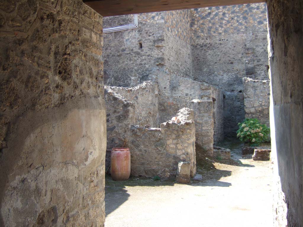 I.10.18 Pompeii. September 2005. Looking south-west from entrance fauces across atrium.
Right of centre is the area of room 6, looking ahead to cistern mouth in walkway No.8.
On the left at the end of the entrance corridor wall is the south-west side of the atrium.
Left of centre is room 7, the kitchen (room 9) is at the rear of room 7.
According to Boyce, in the south wall of the kitchen beside the hearth was a fragmentary painting in two zones.
It was on a white background outlined with red stripes.
In the upper zone stood the Genius, with a Lar.
In the lower zone, beneath garlands, was the tail of a black and yellow serpent.
Below the serpent were several kitchen articles, a hog’s head, a ham on a nail, and an eel on a spit.
See Notizie degli Scavi di Antichità, 1934, p. 344, and fig.38.
See Boyce G. K., 1937. Corpus of the Lararia of Pompeii. Rome: MAAR 14. (p.29, no.60) 
Fröhlich described it was “mostly fallen from the wall, and only the body of the left Lar is quite well preserved.”
See Fröhlich, T., 1991. Lararien und Fassadenbilder in den Vesuvstädten. Mainz: von Zabern. (L18, Taf. 26, 3).