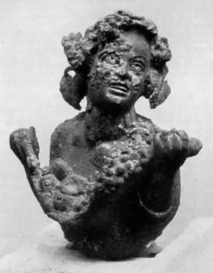 I.16.5 Pompeii. Small bronze bust of smiling satyr.
From the same room as the previous finding, and on the same day, the bronze bust had been found, depicting a satyr holding in his arms, a fleece (?) or a basket full of fruit. 
The satyr turns up his laughing face, slightly leaning towards his left shoulder, the head of bunches of grapes, vine-leaves and branch of ivy make up a strange and lavish hairstyle. 
The small bronze, which originally would have been an artistic part of furniture, probably of the bull (toro?) of a bed or a strong-box, has a maximum height of m.0.095 with a width of m.0.085.
Parco Archeologico di Pompeii. Inventory number 11872. 
It was probably part of all the set of equipment, preserved/stored in the armarium
See Elia, O. Bacco Fanciullo e Dioniso Chtonio a Pompei. Bollettino d'Arte 1961, Fasc. I-II, (p.5, fig.7).