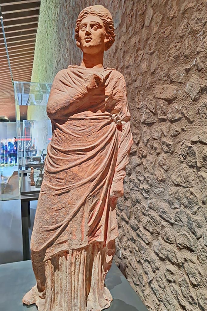 I.20.4 Pompeii. March 2024. Terracotta statue of the muse Polyhymnia.
Found near the north wall of the garden. Traces of red are still preserved on the white folds of the chiton and its mantle.
See Jashemski, W. F., 1993. The Gardens of Pompeii, Volume II: Appendices. New York: Caratzas, p. 67.
At the time of excavation this was recorded as found on 31st January 1959, in I.15.4, which has since been renumbered as I.20.4.
PAP inventory number 12366. Photo courtesy of Giuseppe Ciaramella.
On display in exhibition in Palaestra entitled – “L’altra Pompei, vite comuni all’ombra del Vesuvio”.