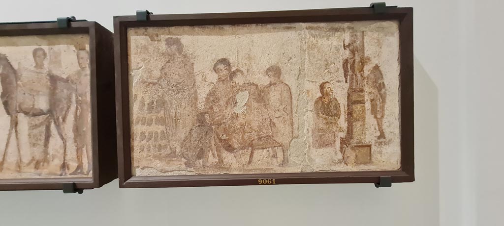 II.4.3 Pompeii. April 2023. Paintings of the Forum frieze, on display in “Campania Romana” gallery in Naples Archaeological Museum.
In centre (inv. 9061) – Vendita di scarpe; scriba e statua equestre. 
(Sale of shoes; scribe and equestrian statue). Photo courtesy of Giuseppe Ciaramella.