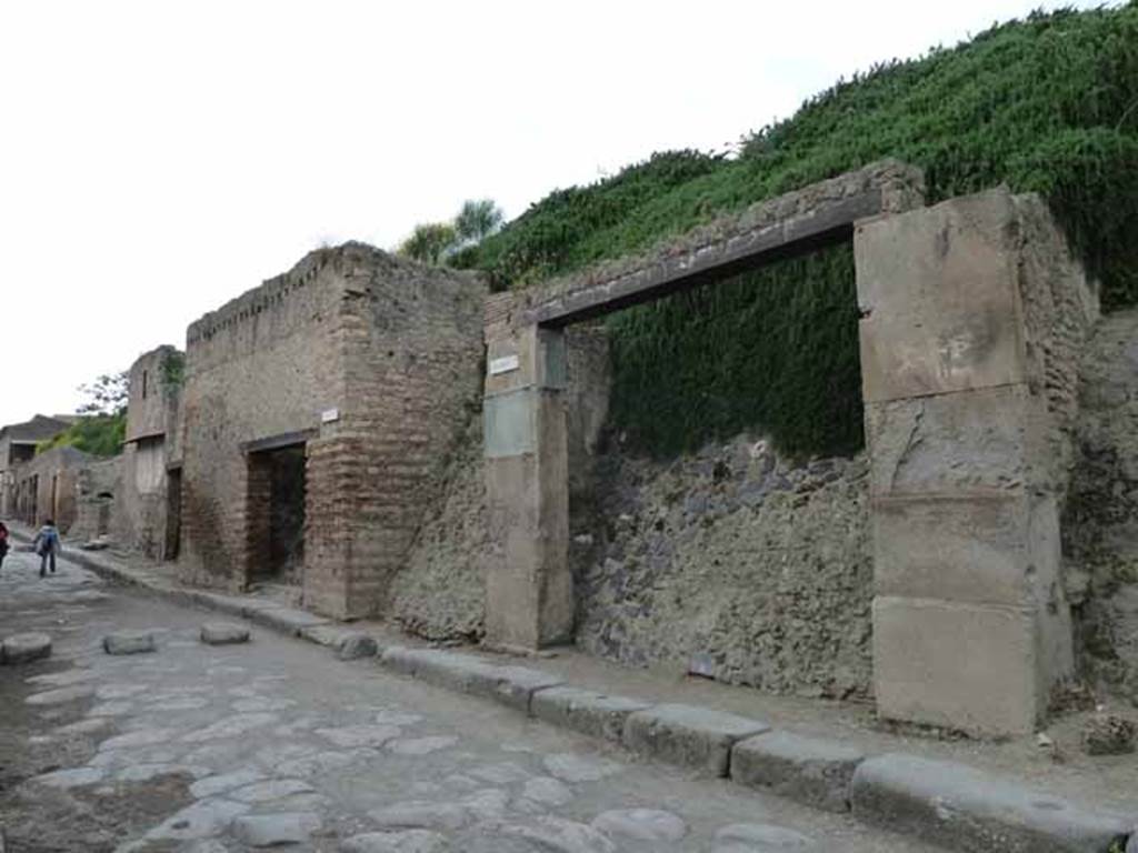 III.3.1 Pompeii. May 2010.  Entrance on Via dell’Abbondanza, looking west past unexcavated roadway to insula III.2.  Originally when excavated in 1914, this doorway was found with carbonised architrave and part of carbonised doorpost.
These were reinstated under glass, and left in situ, after consolidation of the doorway.   See Notizie degli Scavi, 1914, (p.255)
There was also a balcony leaning out over the street, which was reconstituted from its elements,as well as the remains of a stone window frame, to the right (east end).