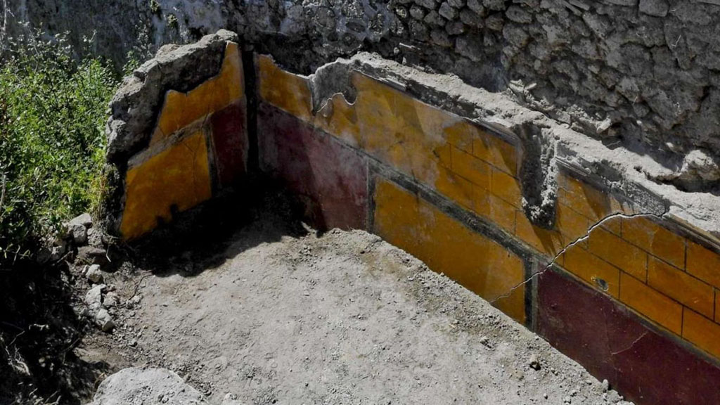 V.2.21 Pompeii. August 2018. Room A9 (as shown on PAP plan), looking towards south and west walls of the cubiculum.
The walls have a cornice at the top, under which are three rows of a yellow painted blockwork design.
Below this are red side panels with a yellow centre panel edged in black.
In the north centre panel is the painting of the wounded Adonis with Venus and cupids.
At the north end of the west and east walls are recesses for a bed.
Photograph © Parco Archeologico di Pompei.