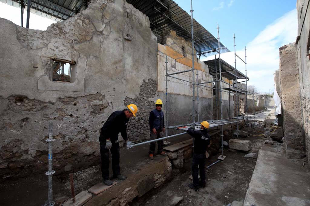 V.2.Pompeii. 
Looking north in Vicolo dei Balconi, with Casa di Orione, entrance doorway A4 on west side, centre right. 
The facade is of white block pattern with a yellow upper band.
Upon excavation, the roadway was discovered still not yet completely paved outside this house, it was presumably still in the course of being re-laid.
At the top of the exterior façade, beam holes for the support of a balcony leaning out over the pavement could be made out.
Photograph © Parco Archeologico di Pompei.
See Osanna M, 2019. Pompei. Il tempo ritrovato. Le Nuove Scoperte. Milano: Rizzoli (p. 89). 
On the north side near to the entrance doorway (centre right of photo), an electoral inscription was painted in red with the name of CELSUM AED O.V.F
This would have been for Lucius Albucius Celsus, followed by AED (aed(ilem) and then O.V.F (o(ro) v(os) f(aciatis), meaning we beg you to vote for Celsus as candidate for aedile.
Above this inscription was another older one painted in red but faded, in larger letters, but unreadable.
See Osanna M, 2019. Pompei. Il tempo ritrovato. Le Nuove Scoperte. Milano: Rizzoli (p. 92).
