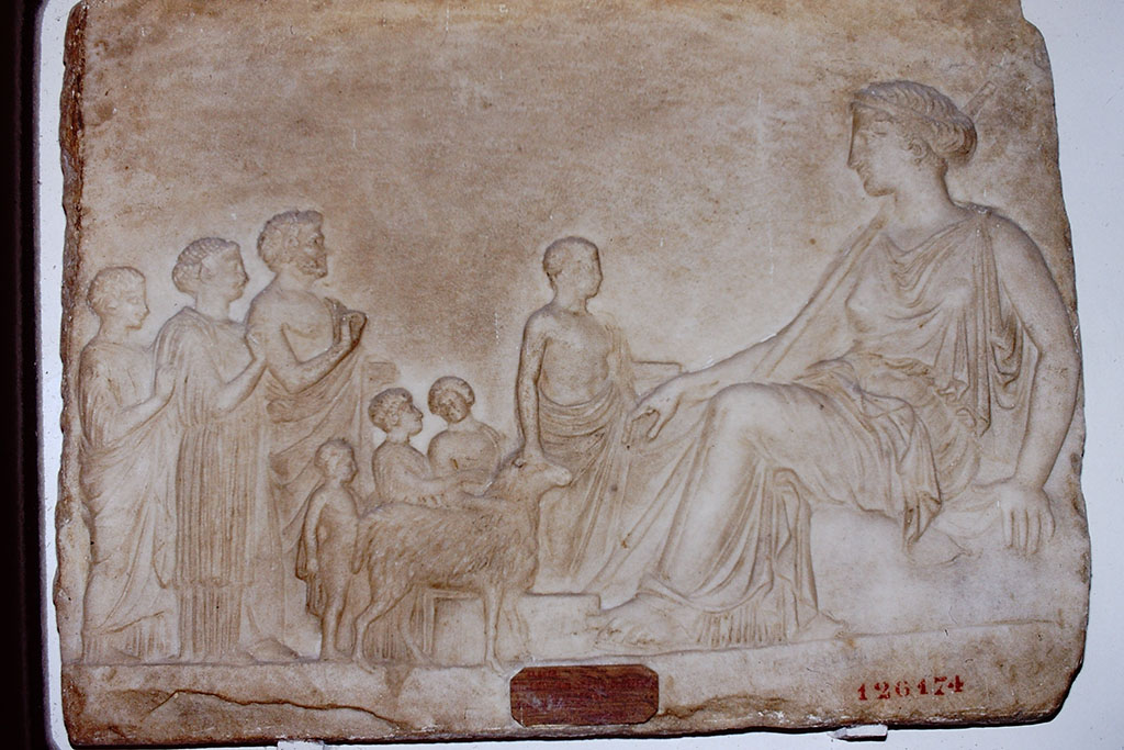 V.3.10 Pompeii. 1984. Marble relief showing a goddess, most likely Aphrodite, enthroned on a rock.
Found on 12th September 1901, in the small garden, not in situ but leaning against the western wall. 
Marble tablet 475mm high, 600mm wide and 45mm thick.
See Notizie degli Scavi di Antichità, 1901, p. 400f.
On the left, in front of the goddess, is a group of two women, a man, three children and an altar servant leading a sacrificial ram.
Now in the Naples Archaeological Museum, inventory number 126174.
According to Kraus, this relief was found leaning against a wall in the small garden, perhaps waiting to be set into the wall. 
See Kraus T. and von Matt L., 1975. Pompeii and Herculaneum: Living cities of the dead. New York: Abrams. (p.193, fig 266
Source: The Wilhelmina and Stanley A. Jashemski archive in the University of Maryland Library, Special Collections (See collection page) and made available under the Creative Commons Attribution-Non Commercial License v.4. See Licence and use details.
J84f0012