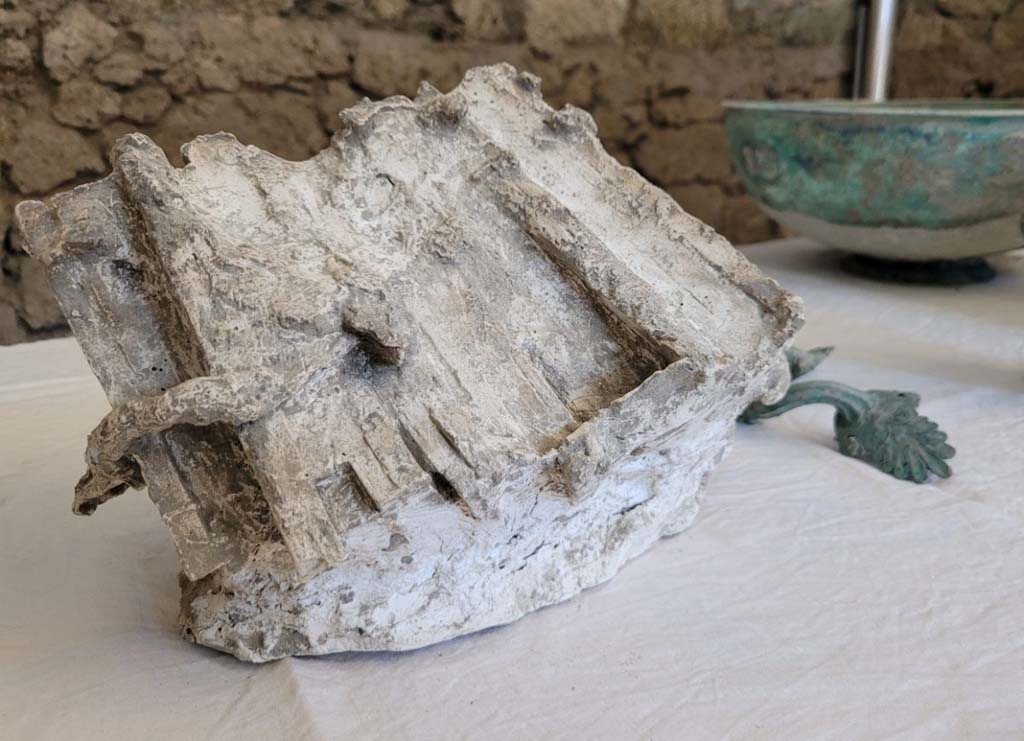 V.3.13 Pompeii. 2022. Upper floor.
The upper rooms were excavated first, however the materials discovered were primarily found where they had fallen into the area of the rooms below.
Amongst these, of great documentary value is the small cast of the waxed tablets. A truly unique find, it has allowed us to create the first example of a cast which allows a perfect yield of both volume and detail. It is a group of seven triptychs, tied together by a small cord both horizontally and vertically. The polyptych had probably been stored on a shelf, together with other ceramic and bronze objects.
Discovered within a large cupboard which collapsed during the eruption were various other well-preserved ceramic vessels for everyday use in the kitchen and the canteen, but also vessels in sigillata (a type of Roman fine table ceramic) and in glass. Alongside this was a set of bronze vessels, among which a well-preserved pelvis (bowl) with a beaded base and handles with palmette attachments particularly stands out. There are in addition two bronze jugs, one of which features a handle with a sphinx-shaped attachment above and a lion’s head attachment below.
Gli ambienti superiori sono stati scavati per prima, tuttavia i materiali rinvenuti sono stati prevalentemente ritrovati in fase di caduta nella volumetria degli ambienti sottostanti. 
Tra questi di gran valore documentario è il piccolo calco delle tavolette cerate. Un unicum per la tipologia di ritrovamento, che ne ha permesso di realizzare il primo esemplare di calco, che ne consente la perfetta restituzione della volumetria e dei dettagli. Si tratta di un gruppo di sette trittici, legati tra essi da un cordino sia in senso orizzontale che verticale. Il polittico doveva probabilmente essere conservato su qualche scaffale, unitamente ad altri oggetti in ceramica e in bronzo. 
Contenute all’interno di un grosso armadio, crollato durante l’eruzione, sono inoltre state recuperate diverse forme ceramiche d’uso comune, da cucina e da mensa, ma anche forme in sigillata (tipo di ceramica romana fine da mensa) ed in vetro, molto ben conservate. A queste si affianca un piccolo set di forme in bronzo, tra cui spicca una ben conservata pelvis (bacile) con fondo perlinato ed anse con attacchi a palmette. Con essa anche due brocche bronzee, una delle quali con ansa con applique sormontante a forma di sfinge ed attacco inferiore a testa leonina. 
Photograph © Parco Archeologico di Pompei.