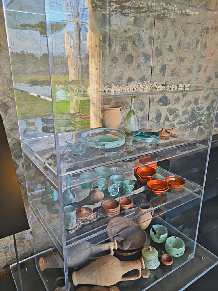 V.3.13 Pompeii. March 2024. 
Reconstruction of a cupboard/cabinet (originally made of wood) with its original contents, found in this house. There was a large amount of crockery mainly pottery and glass. On shelves 3 and 4 are the remains of carbonised wooden planks. 
Ricostruzione di un armadio/mobile (originariamente in legno) con il suo contenuto originale, trovato in questa casa. C'era una grande quantità di stoviglie, principalmente ceramiche e vetro. Sugli scaffali 3 e 4 ci sono i resti di assi di legno carbonizzato.
On display in Palaestra in exhibition entitled – “L’altra Pompei, vite comuni all’ombra del Vesuvio”.
Photo courtesy of Giuseppe Ciaramella.