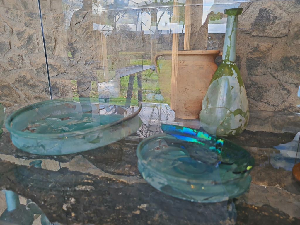 V.3.13 Pompeii. March 2024. Restored glass items and carbonised wooden shelf in reconstruction of a cupboard/cabinet. 
Oggetti in vetro restaurati e mensola in legno carbonizzato nella ricostruzione di un armadio/armadio.
Photo courtesy of Giuseppe Ciaramella.
On display in exhibition in Palaestra entitled “L’altra Pompei, vite comuni all’ombra del Vesuvio”.