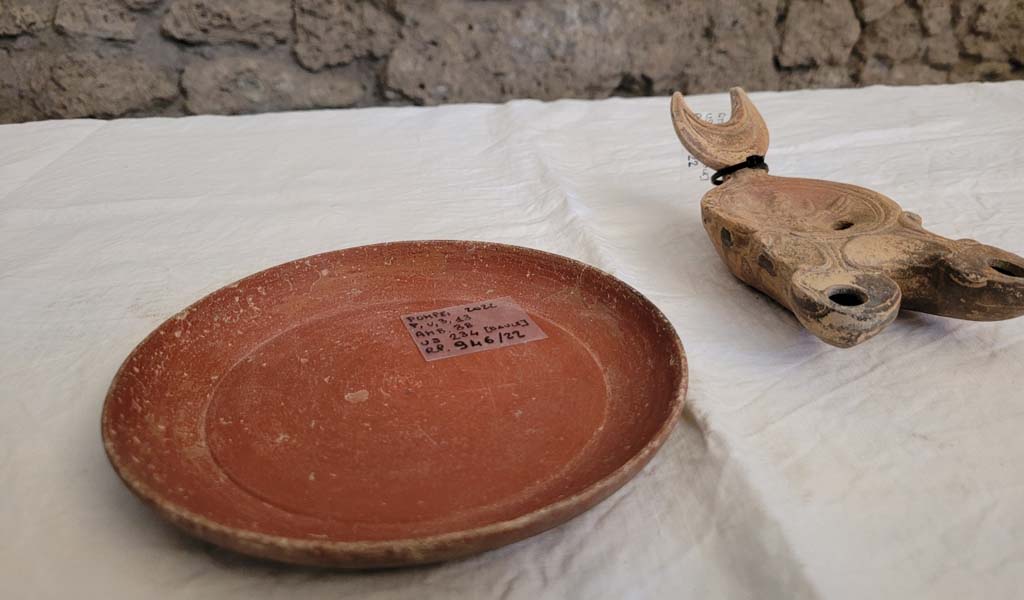 V.3.13 Pompeii. 2022. Room 3b, bedroom. Chest contents.
A small sigillata plate and a double-spouted lantern with a bas-relief depiction of the transformation of Zeus into an eagle.
Un piattino in sigillata (tipo di ceramica romana fine da mensa) e una lucerna a doppio beccuccio con bassorilievo raffigurante la trasformazione di Zeus in aquila. 
Photograph © Parco Archeologico di Pompei.