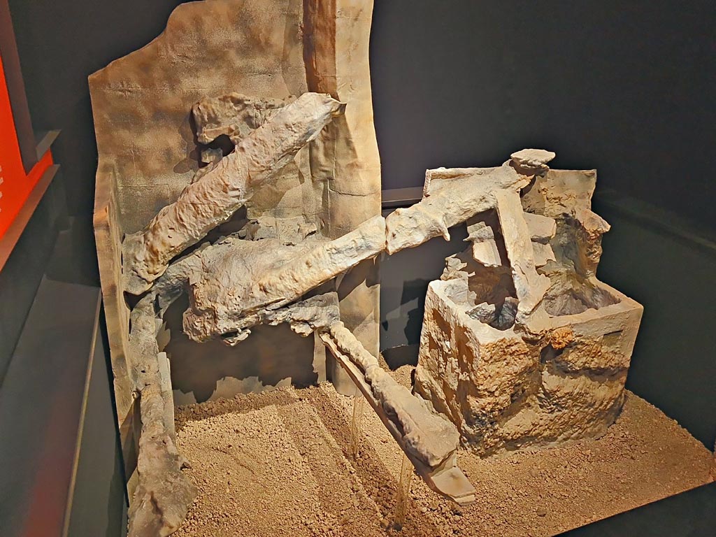 V.3.13 Pompeii. March 2024. 
Room 3b, on the left is the cast of the remains of a bed with a headboard to which a pillow is attached.
On the right is a copy of an open wooden trunk, on which the beams and boards of the floor above have collapsed.
Next to it, is a round three-legged table (see photo above this one).  
On display in exhibition in Palaestra entitled “L’altra Pompei, vite comuni all’ombra del Vesuvio”. Photo courtesy of Giuseppe Ciaramella.