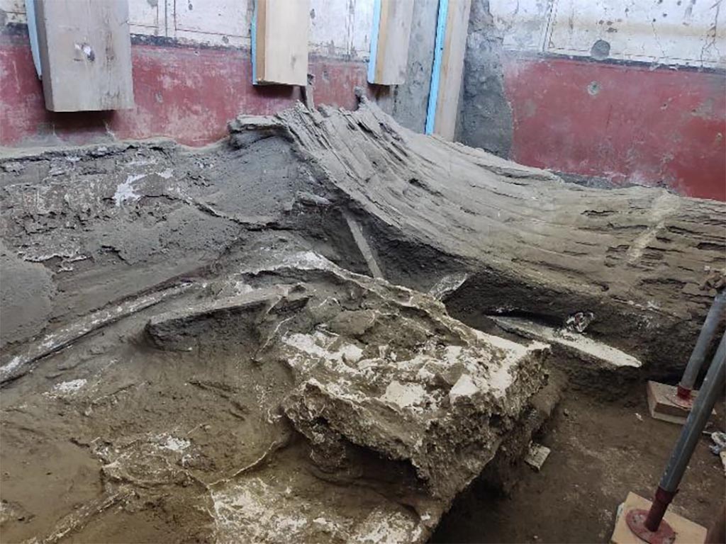 V.3.14 Pompeii. August 2022. Room 16. Letto 1 e letto 2.
At the foot of the first bed, the cast of a probable blanket.
Above the second bed, part of the collapse of the suspended ceiling therefore remains. Here too, through the technique of casts, it was possible to recover the detailed volume of the lath contained in the heart of the mortar of the false ceiling, succeeding in identifying the various bundles composed of a dozen or so thin straws, tied together with a strap and evidently covered with a gauze, probably covered with pitch, that insulated the straws from the damp mortar in which they were sunk. It represents one of the very few material examples of this technique for lightening ceilings, known mainly from Vitruvius, De architectura... VII, 3.
At the foot of the second bed, near the south-west corner, the cinerite has preserved the trace of the bottom of a wicker basket of just over 30 cm in diameter as well as a small quadrangular storage unit of approx. 50 x 45 cm in size. The execution of the casts of these last two pieces of furniture has been arranged in the last few days.
Ai piedi del primo letto, il calco di una probabile coperta.
Al di sopra di questo secondo letto, insiste dunque parte del crollo del controsoffitto. Anche qui attraverso la tecnica dei calchi, è stato possibile recuperare il volume dettagliato dell’incannucciata contenuta nel cuore della malta del controsoffitto, riuscendo ad identificare i diversi fasci composti da una decina, o poco più, di sottili cannucce, legate tra loro con un laccetto ed evidentemente rivestite da una garza, probabilmente impeciata, che isolava le cannucce dalla malta umida in cui affondavano. Esso rappresenta uno dei pochissimi esempi materiali di tale tecnica di alleggerimento dei solai, nota principalmente da Vitruvio, De architectura… VII, 3.
Ai piedi del secondo letto, presso l’angolo sud-ovest, la cinerite ha conservato la traccia del fondo di una cesta in vimini di poco più di 30 cm di diametro nonché di un piccolo mobile contenitore quadrangolare di ca. 50 x 45 cm. Di questi ultimi due elementi d’arredo sono stati disposti in questi giorni l’esecuzione dei calchi.
Photograph © Parco Archeologico di Pompei.