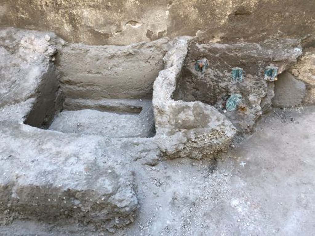 V.3 Pompeii. Casa del Giardino. August 2019. Outline of the wooden box which contained the gems.
Kept in a wooden case, and recently restored and brought to their former glory by the restorers of the Restoration Laboratory of the Archaeological Park of Pompeii, they formed part of the precious valuables of the family, which perhaps the inhabitants of the house could not take away before attempt ing to escape. The trace of the wooden case that contained the finds, whose bronze hinges have been well preserved inside the volcanic material, unlike the wooden decomposed part, was identified next to the imprint of another case or piece of furniture in the corner of one of the service areas, probably used as a repository.
Impressione della cassa di legno che conteneva le gemme.
Custoditi in una cassa in legno, e da poco restaurati e riportati al loro splendore dalle restauratrici del Laboratorio di Restauro del Parco Archeologico di Pompei, si trattava di una parte dei preziosi di famiglia, che forse gli abitanti della casa non riuscirono a portare via prima di tentare la fuga. La traccia della cassa in legno che conteneva i reperti, le cui cerniere bronzee si sono ben conservate all’interno del materiale vulcanico, a differenza della parte lignea decompostasi, è stata individuata accanto all’impronta di un’altra cassa o mobile nell’angolo di uno degli ambienti di servizio, probabilmente usato come deposito.
Photograph © Parco Archeologico di Pompei.