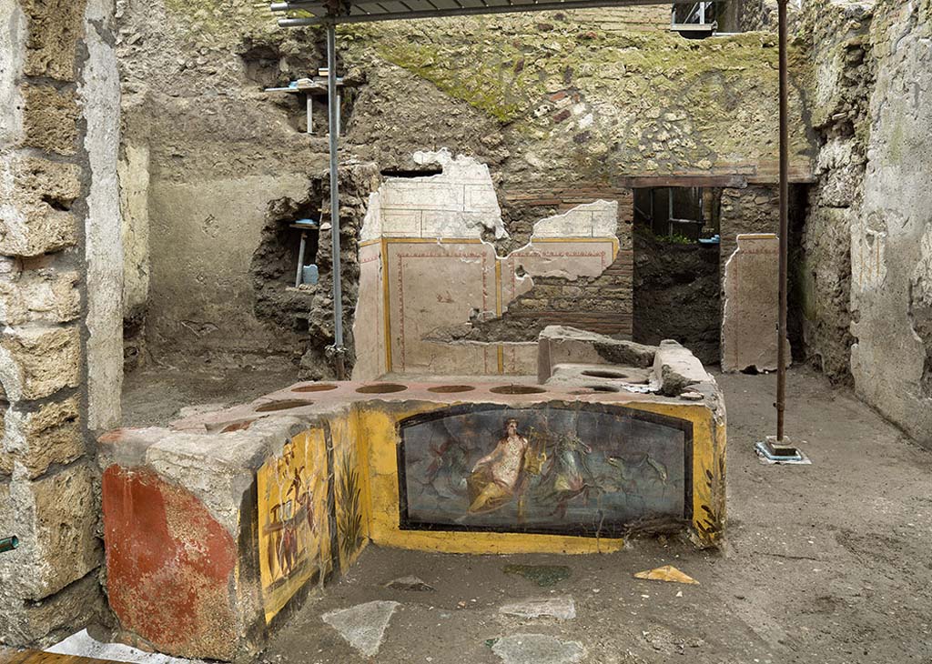 V.3 Pompeii, Thermopolium with painting of nereid. December 2020. Looking across the counter and rooms from the front entrance on the square in front of Vicolo delle Nozze d'Argento.
The flooring of the entire room consisted of a layer of cocciopesto (a waterproof covering made of terracotta fragments), into which fragments of polychrome marble (alabaster, portasanta, green breccia and bardiglio) were inserted in several areas.
V.3 Pompei, Termopolio con dipinto di nereide e anfore. Dicembre 2020. Guardando dall'altra parte del bancone e le camere dall'ingresso anteriore sulla piazza di fronte a Vicolo delle Nozze d'Argento.
Il piano pavimentale di tutto l'ambiente è costituito da uno strato di cocciopesto (rivestimento impermeabile composto da frammenti in terracotta), in cui in alcuni punti sono stati inseriti frammenti di marmi policromi (alabastro, portasanta, breccia verde e bardiglio).
Photo by Luigi Spina courtesy of Parco Archeologico di Pompei.