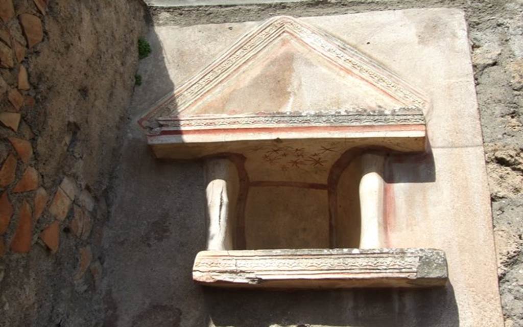 V.4.9 Pompeii. March 2009. Lararium niche. According to Boyce, this Lararium was on a special panel of rough white stucco, and was adorned with an aedicula façade. Beneath the niche was a heavy shelf with two columns resting on it, framing either side of the niche. The interior of the niche was white and outlined with red stripes. On the ceiling of the niche there were red and green star decorations. Within the niche were found the following figures – A bronze bust of Minerva, wearing the aegis and helmet. She was leaning backwards in a strange manner upon a bronze plate fitted with a sort of handle at the back.
A roughly worked alabaster figure of Venus wearing a crown, beside her to the left stands a small figure clad in a long garment. Venus seems to held her left hand around the smaller figure, while the latter placed her right hand upon the head of the goddess. This group stood upon a marble-covered base behind the bust of Minerva. A small standing male figure of ivory. Sogliano thought the figure was Venus, Mau thought it was definitely male.
Also found in the niche were two small rectangular terracotta altars and two terracotta plates. See Notizie degli Scavi di Antichità, 1899,pp.103. See Boyce G. K., 1937. Corpus of the Lararia of Pompeii. Rome: MAAR 14. (p.41, no.123, Pl.7,1).