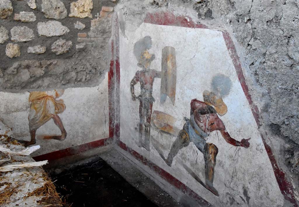 The fresco of about 1.12m x 1.5m, found in an environment behind the wide intersection of the Vicolo dei Balconi and the Vicolo delle Nozze d’Argento, has the shape of a trapezoid, as it was placed under the stairs, perhaps of a workshop. Above the painting, the imprint of the wooden staircase can be seen above the painting.  Most likely it decorated an environment frequented by gladiators, perhaps a bar equipped with an upper floor, intended for the accommodation of the owners of the business or as frequently, especially given the presence of gladiators, intended for prostitutes.
L’affresco di circa 1,12 mt x 1,5mt, rinvenuto in un ambiente alle spalle dello slargo di incrocio tra il Vicolo dei Balconi e il vicolo delle Nozze d’Argento, ha forma trapezoidale, in quanto era collocato nel sottoscala, forse di una bottega. Si intravede al di sopra della pittura, l’impronta della scala lignea.  Molto probabilmente decorava un ambiente frequentato da gladiatori, forse una bettola dotata di un piano superiore, destinato ad alloggio dei proprietari dell’esercizio commerciale o come di frequente, soprattutto vista la presenza di gladiatori, destinato alle prostitute.
Photograph © Parco Archeologico di Pompei.
Vedi http://pompeiisites.org/comunicati/laffresco-dei-gladiatori-combattenti-nuova-scoperta-nella-regio-v/