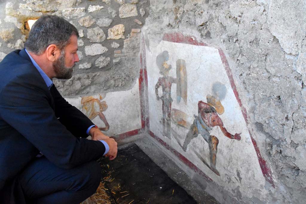 Massimo Osanna studies the fresco of two gladiators at the end of the fight, one wins the other succumbs.  
It is the scene of the latest fresco found in Pompeii in Regio V, as part of the work of securing and remodulating the excavation fronts, provided for by the Great Pompeii Project. It is the scene of a fight between a "Mirmillo" and a "Thracian", two types of fighters distinct from different armour and classic opponents in gladiatorial fights. The discovery environment is only partially brought to light - on one side emerges another small portion of fresco that reveals the presence of another figure.
Massimo Osanna studia l'affresco di due gladiatori al termine del combattimento, l’uno vince l’altro soccombe. E’ la scena dell’ultimo affresco rinvenuto a Pompei nell’area di cantiere della Regio V, nell’ambito dei lavori di messa in sicurezza e rimodulazione dei fronti di scavo, previsti dal Grande Progetto Pompei. Si tratta della scena di un combattimento tra un “Mirmillone” e un “Trace”, due tipologie di lottatori distinti da armature differenti e classici avversari nelle lotte gladiatorie.
L’ambiente di rinvenimento è solo parzialmente portato in luce - su un lato emerge un'altra piccola porzione di affresco che rivela la presenza di un’altra figura.
Photograph © Parco Archeologico di Pompei.