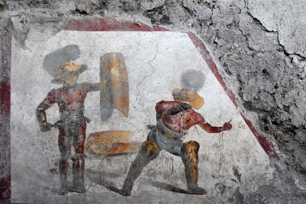 A contest between a Mirmillo and a "Thracian", two types of fighters distinct from different armour and classic opponents in gladiatorial fights. 
On a white background, bordered on three sides by a red band, the scene of struggle between the two gladiators is composed. 
On the left is a "Mirmillo" of the "Scutati" category: he wields the offensive weapon, the gladium, (short Roman sword) and a large rectangular shield (scutum) and wears a wide-brimmed helmet equipped with visor with plumes, the cimian. 
The other, in a succumbing position, is a "Thracian", a gladiator of the "Parmularii" category, with the shield on the ground. 
He is represented with a helmet (galea), with wide brim and wide visor to protect the face, topped by a tall crest.
Si tratta della scena di un combattimento tra un “Mirmillone” e un “Trace”, due tipologie di lottatori distinti da armature differenti e classici avversari nelle lotte gladiatorie.
Su uno sfondo bianco, delimitato su tre lati da una fascia rossa, si compone la scena di lotta tra i due gladiatori. Quello sulla sinistra è un “Mirmillone” della categoria degli "Scutati”: impugna l’arma di offesa, il gladium, (corta spada romana) e un grande scudo rettangolare (scutum) e veste un elmo a tesa larga dotato di visiera con pennacchi, il cimiero. L’altro, in posizione soccombente, è un “Trace”, gladiatore della categoria dei "Parmularii”, con lo scudo a terra. È rappresentato con elmo (galea), a tesa larga ed ampia visiera a protezione del volto, sormontato da un alto cimiero.
Photograph © Parco Archeologico di Pompei.