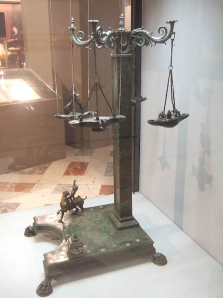 Found 19th August 1813 in VI.6.1 Pompeii. Bronze candelabrum with four volute branches for lamps. 
Now in Naples Archaeological Museum. Inventory number 4563. 
According to Pagano and Prisciandaro, an elegant bronze lamp was found in the portico of the garden.
See Prisciandaro, R., 2006. Studio sulle provenienze degli oggetti rinvenuti negli scavi borbonici del regno di Napoli. Naples: Nicola Longobardi. 
(p. 109, reported 9th September 1813, PAH I, 3, 127, add. 271, and p.109, reported 23rd December 1813, PAH I, 3, 139, add. 271,72).
According to Kuivalainen, this lampstand was discovered in the portico of the peristyle together with the bronze statue of two males, H13 see below).
He comments –
“This lamp has both a religious and a practical purpose. The pictorial programme of the lamp is quite obviously dedicated to Bacchus – the child riding a panther, the mask, the bull as one of his characteristic animals, all speak for this. Choosing a child Bacchus was relatively original due to his rare appearance in visual arts in general, but it may share the idea of growth and fertility with a rhyton, a she-panther, not to omit the idea of light in the worship of Bacchus. Perhaps this lampstand was used for private worship. On the practical side, the asymmetrical placement of the pillar made more space for a vessel with oil for filling the lamps.”
See Kuivalainen, I., 2021. The Portrayal of Pompeian Bacchus. Commentationes Humanarum Litterarum 140. Helsinki: Finnish Society of Sciences and Letters, (H18, p.222).