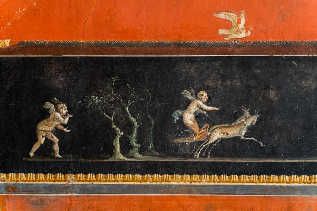VI.15.1 Pompeii. March 2023.
Lower centre of east wall, detail of part of painted panel with cupids in a chariot race. Photo courtesy of Johannes Eber.